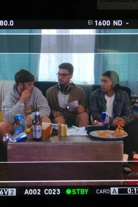 Three young men sitting at a table, one with headphones around his neck, surrounded by snacks and drinks, in a room with a window and curtains in the background.
