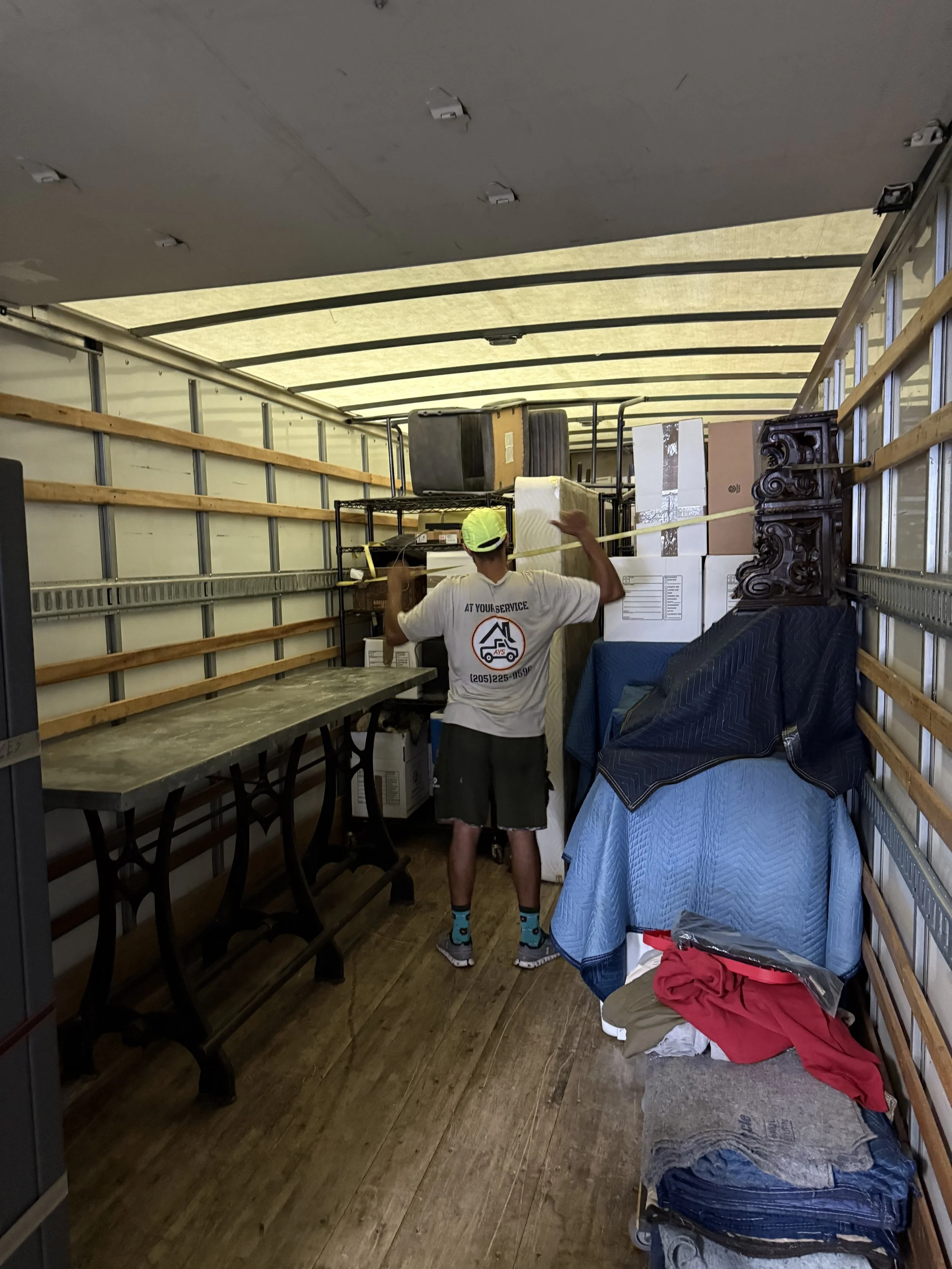 A person wearing a white shirt, shorts, and teal socks is standing inside the back of a moving truck, marking or measuring the stacked items with yellow tape. The truck contains stacked boxes, furniture, and miscellaneous household items.