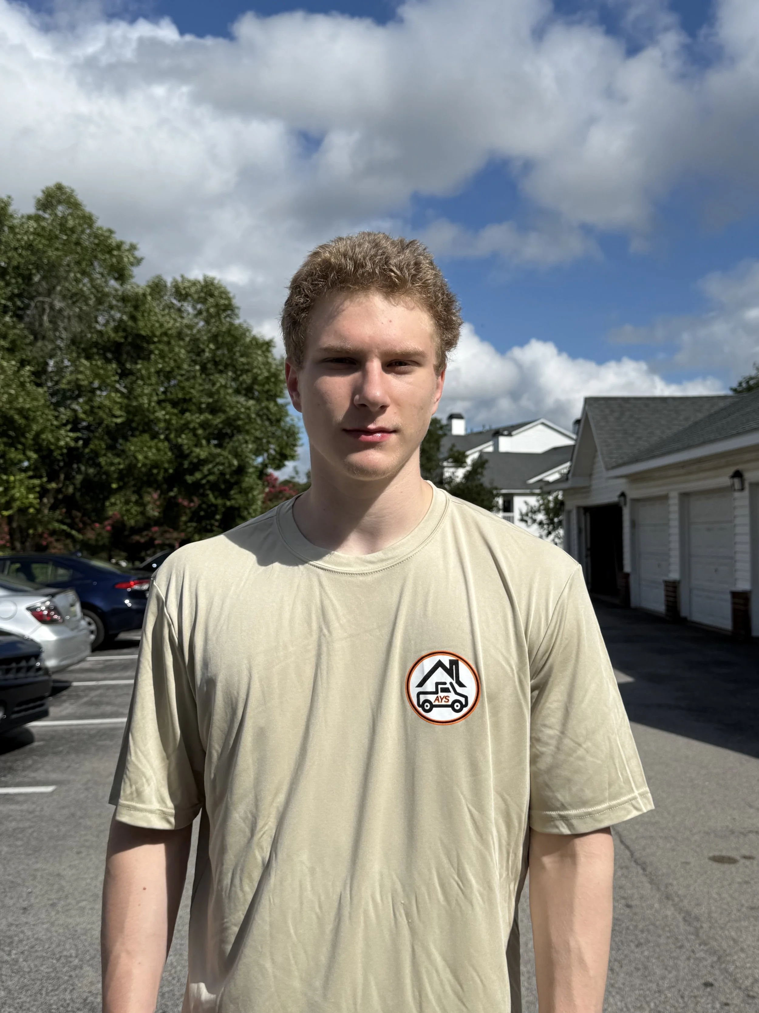 A young man with curly blond hair standing outdoors in a parking lot on a sunny day with partly cloudy skies, wearing a beige T-shirt with a logo on the chest.