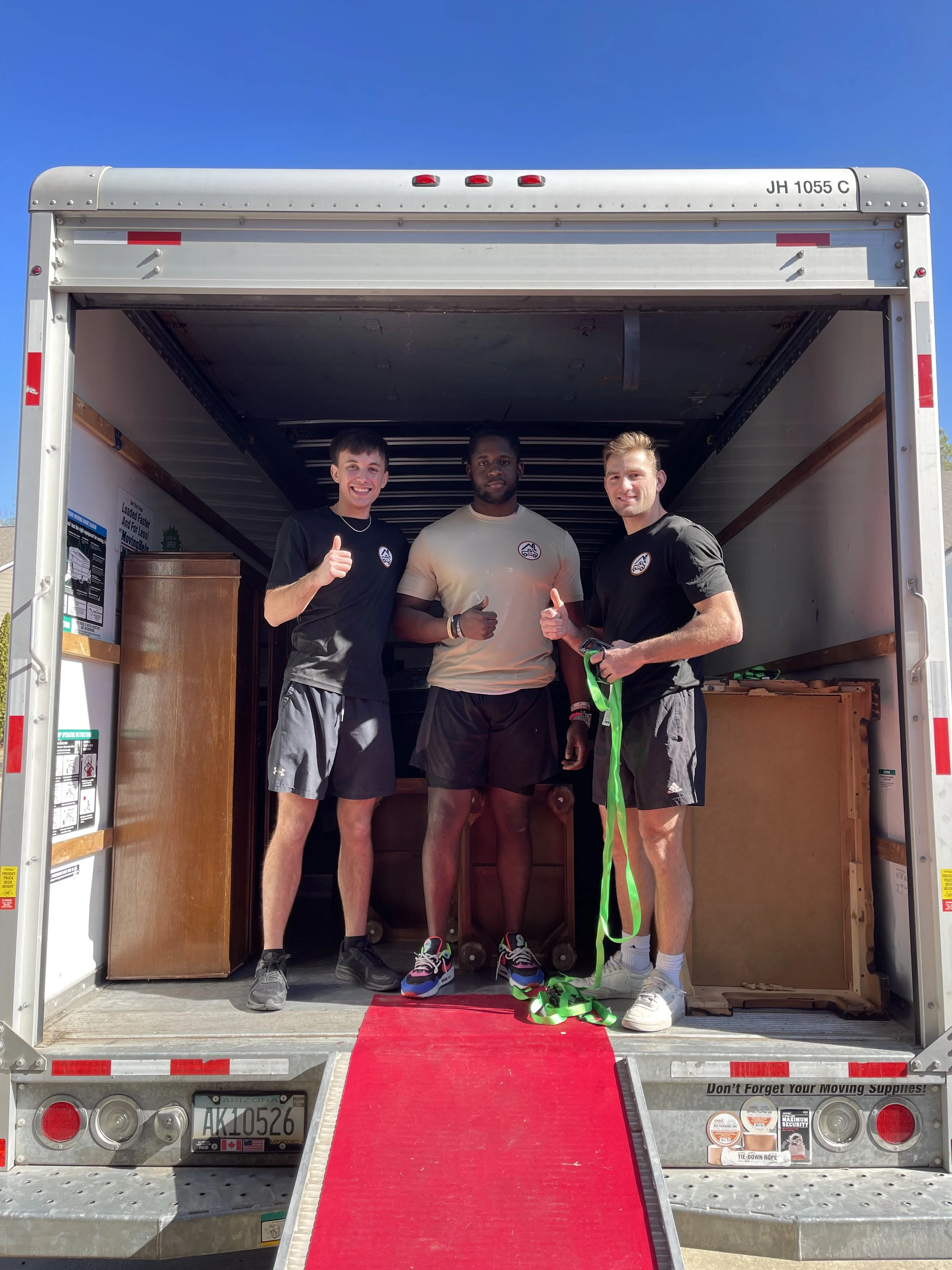 Three young men standing inside a moving truck, smiling and giving thumbs-up signs, with a red moving ramp leading into the truck.