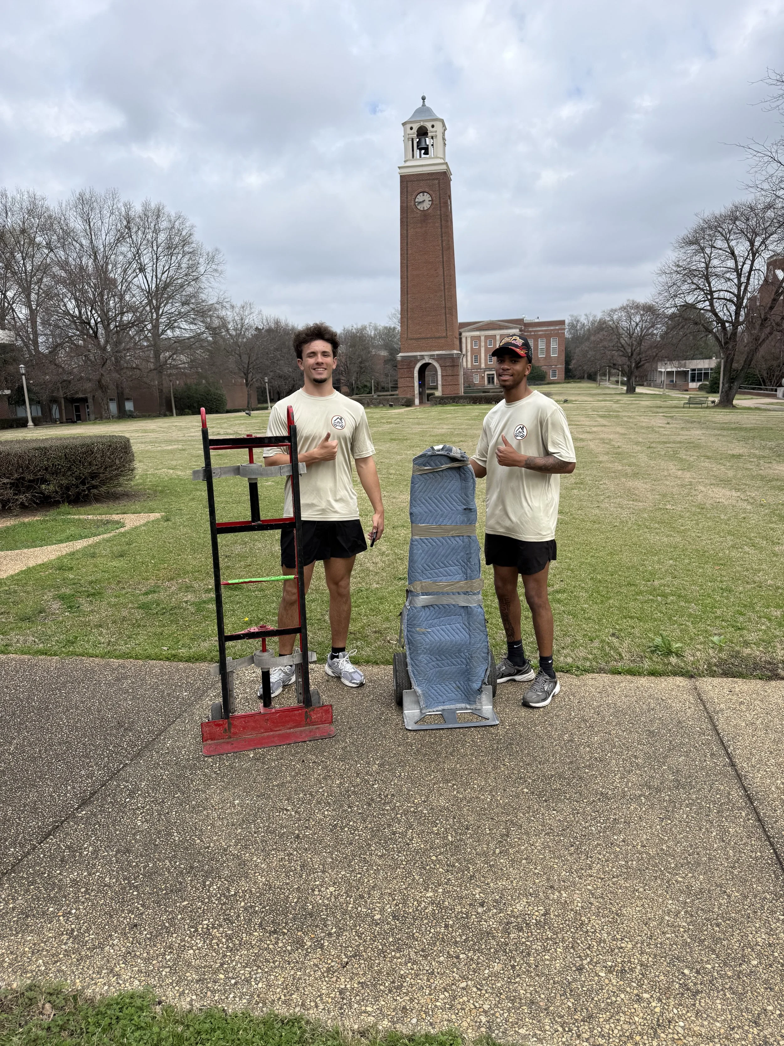 Two young men standing outdoors on a cloudy day, giving thumbs up, with equipment including a red and black ladder and a blue cart, in front of a grassy park area with a tall clock tower and trees in the background.