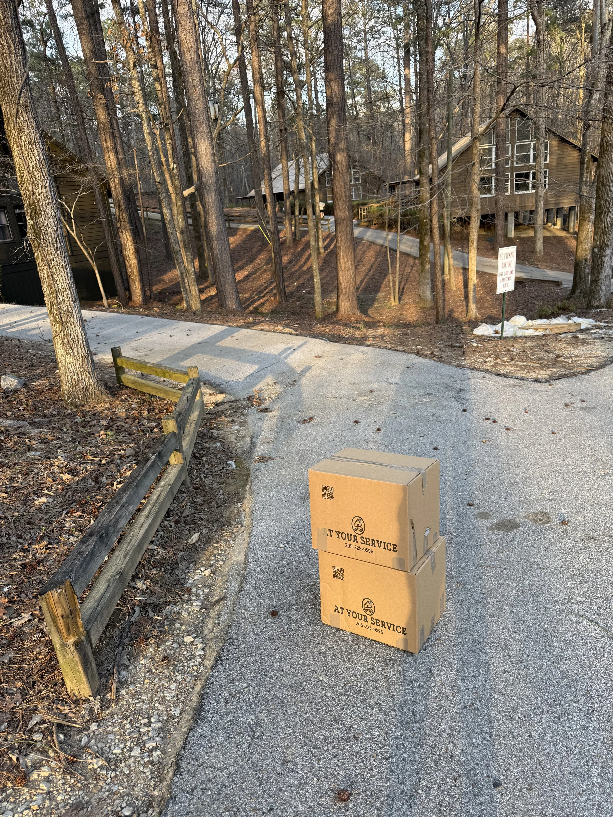 Two stacked cardboard boxes labeled "AT YOUR SERVICE" with a phone number, placed on a paved pathway in a wooded area. In the background, there are trees, residential buildings, a wooden fence, and a sign.
