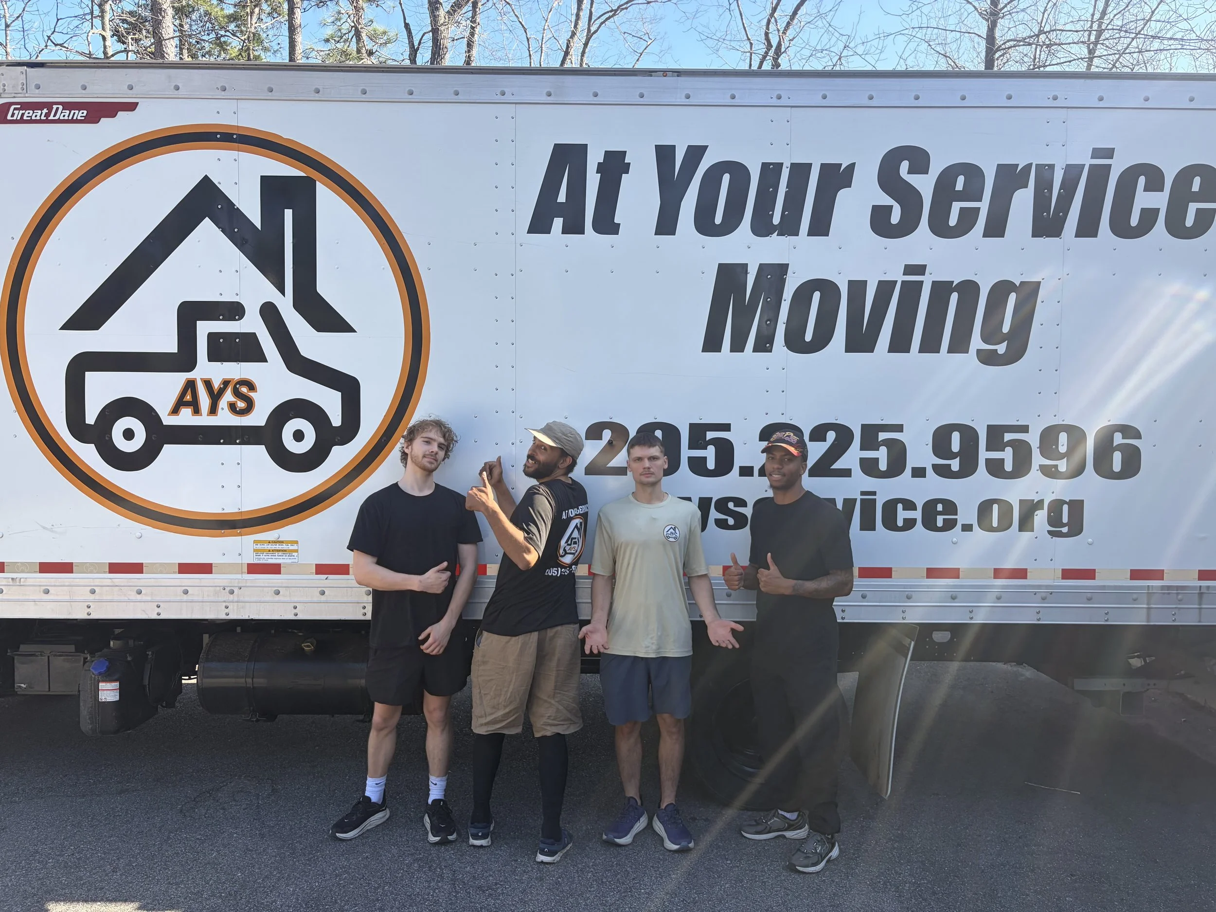 Four people standing in front of a moving truck with a logo for AYS and a house and car graphic, and the text 'At Your Service Moving' along with contact information.