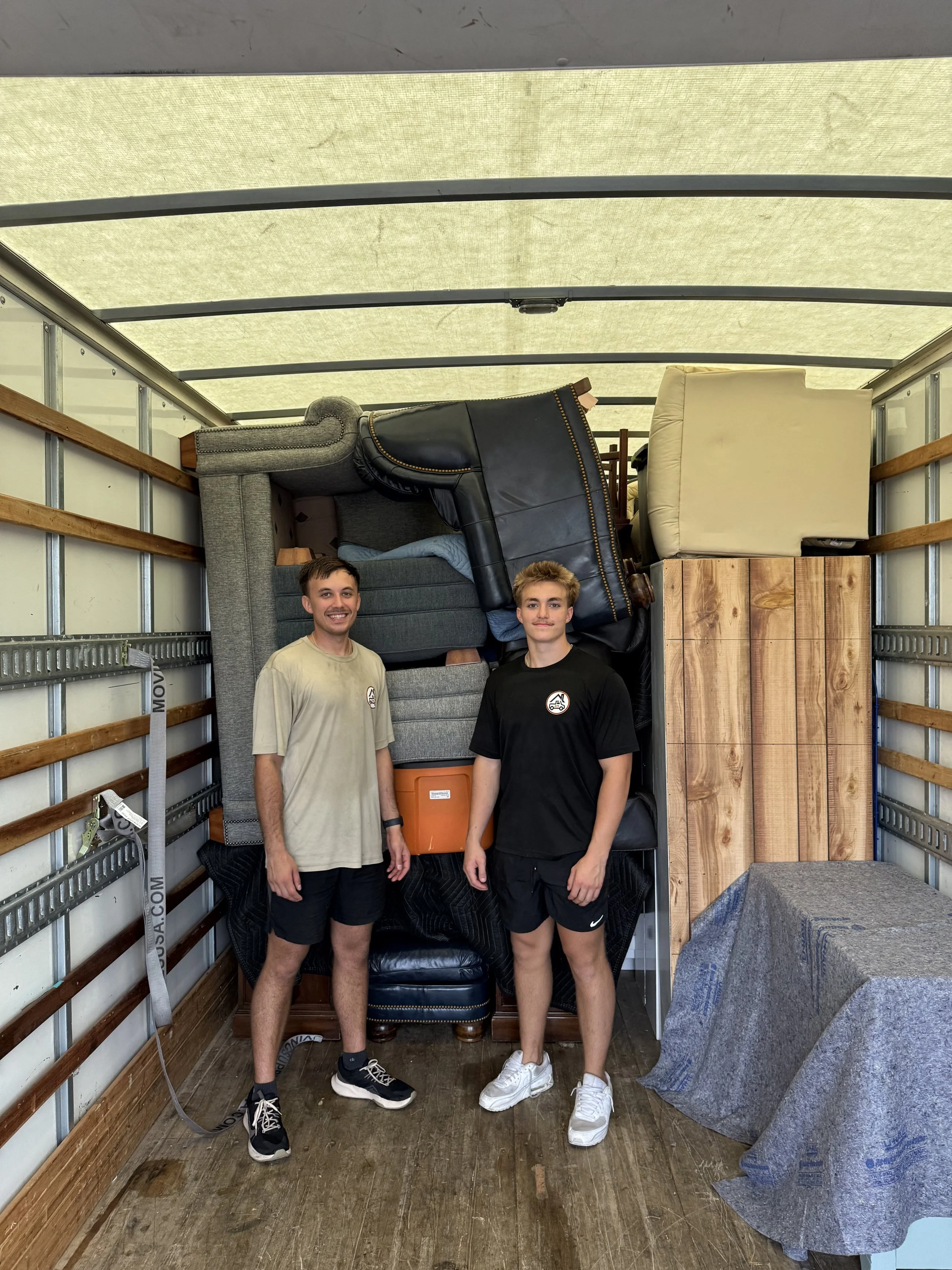 Two young men standing inside a moving truck with furniture piled up behind them.