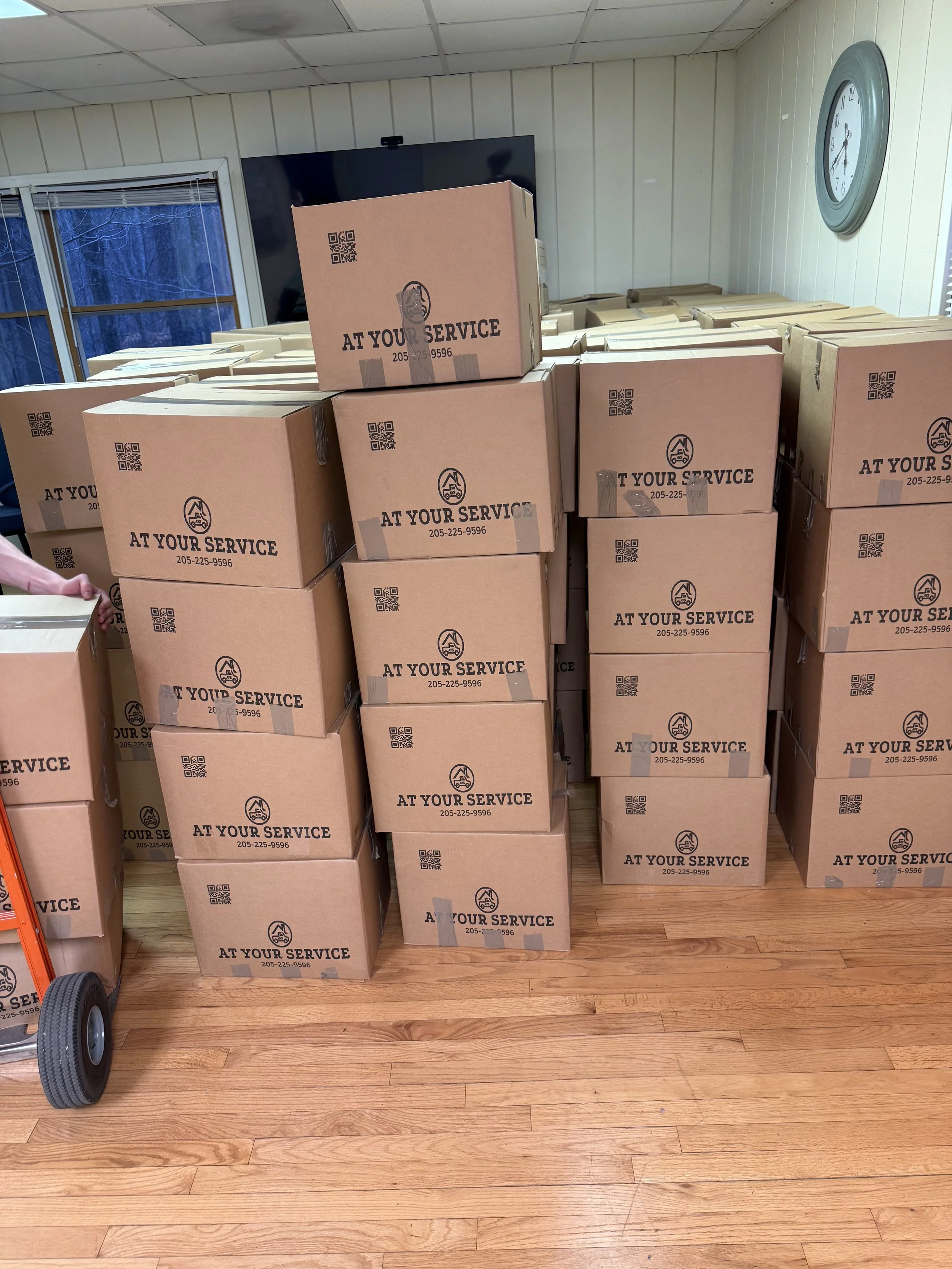 Stacks of cardboard boxes labeled "At Your Service" with a phone number, in a room with hardwood floors and a wall clock.