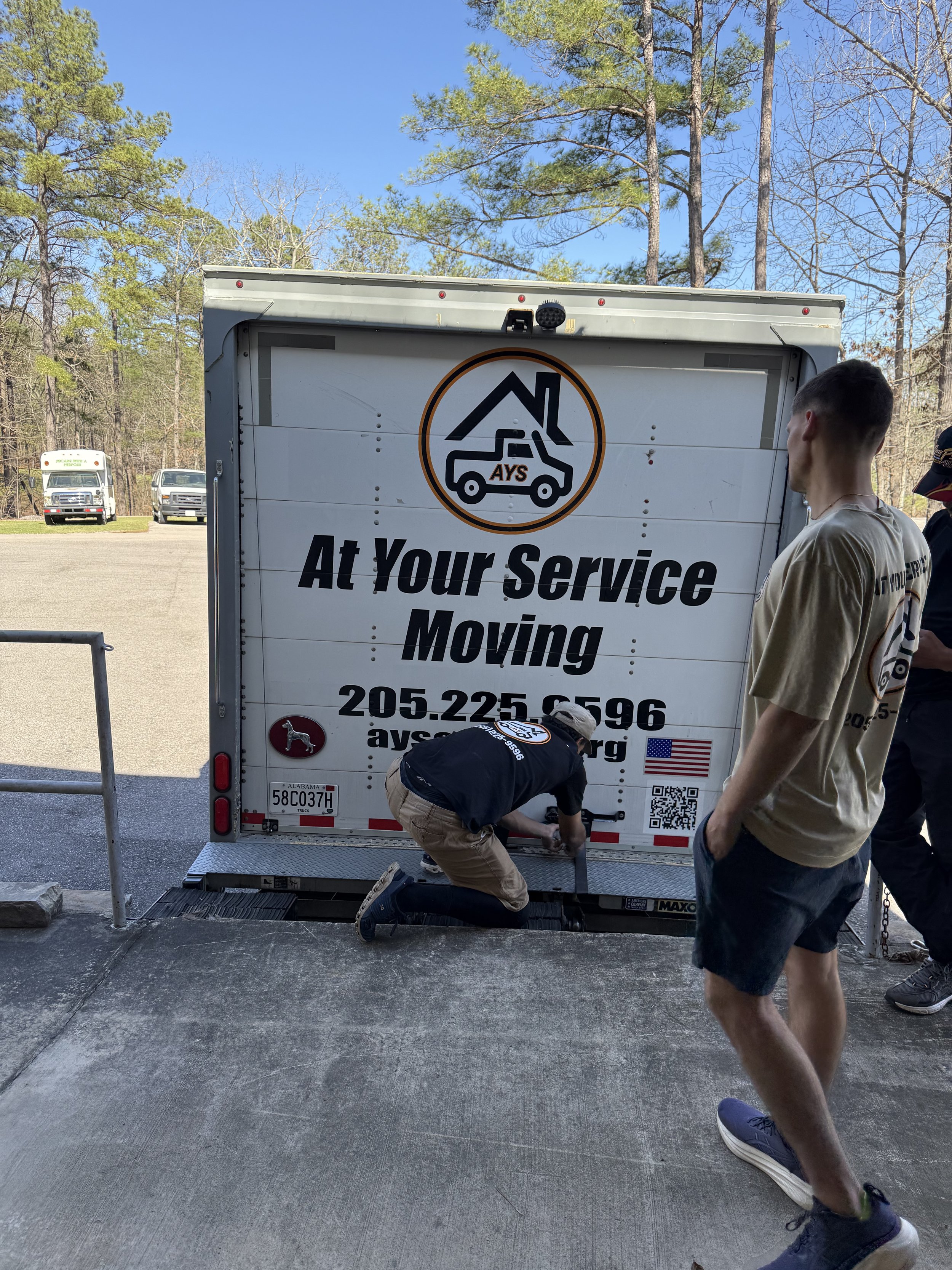 Three people are working around a moving truck with the text 'At Your Service Moving' and contact information. One person is kneeling and securing the truck, while the other two stand nearby.