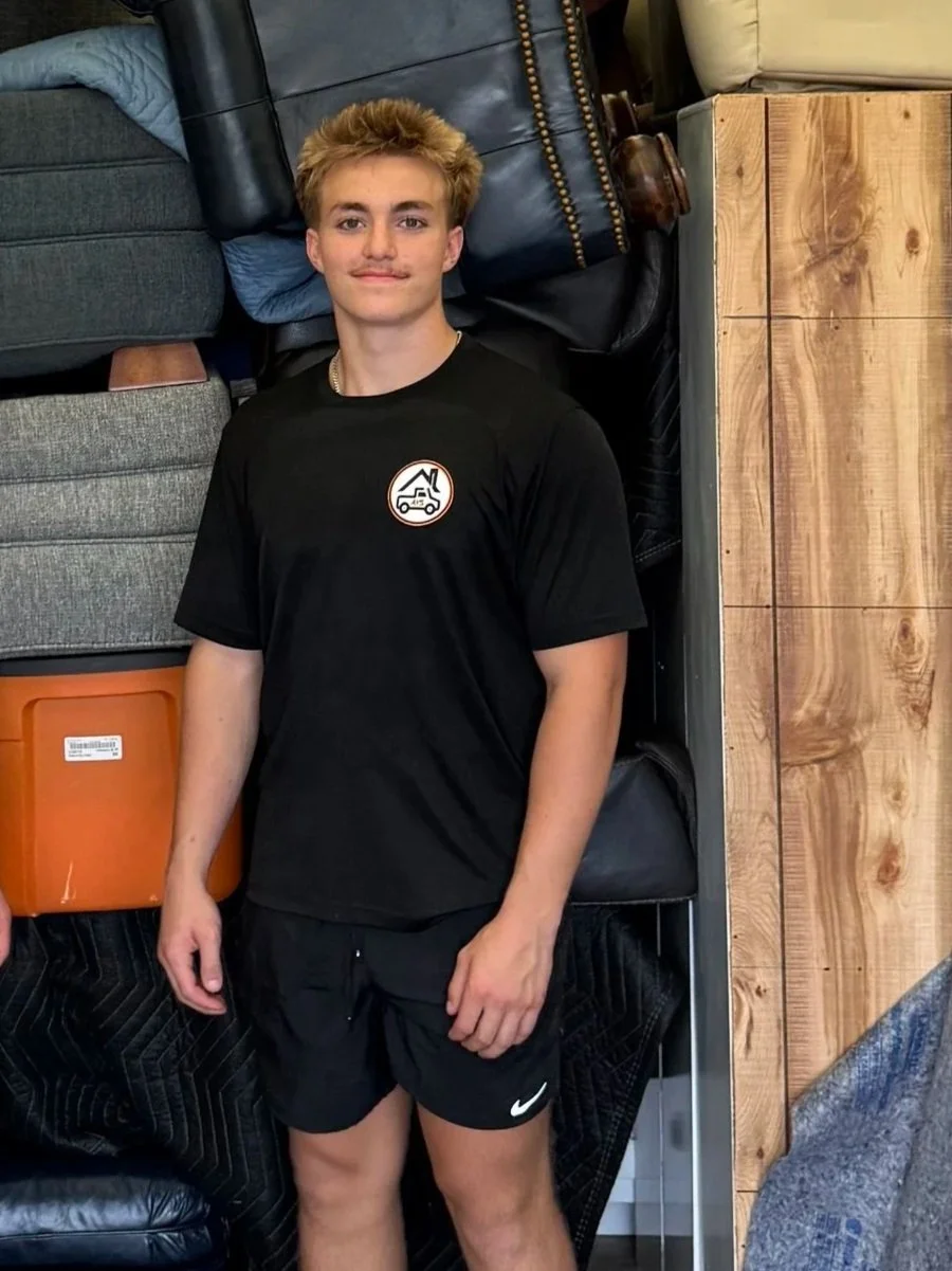 A young man with light hair and a slight mustache is standing indoors, leaning against a variety of furniture including couches and a wooden crate. He is wearing a black T-shirt with a circular logo featuring a mountain, a tent, and a vehicle, and black shorts with a white Nike logo.