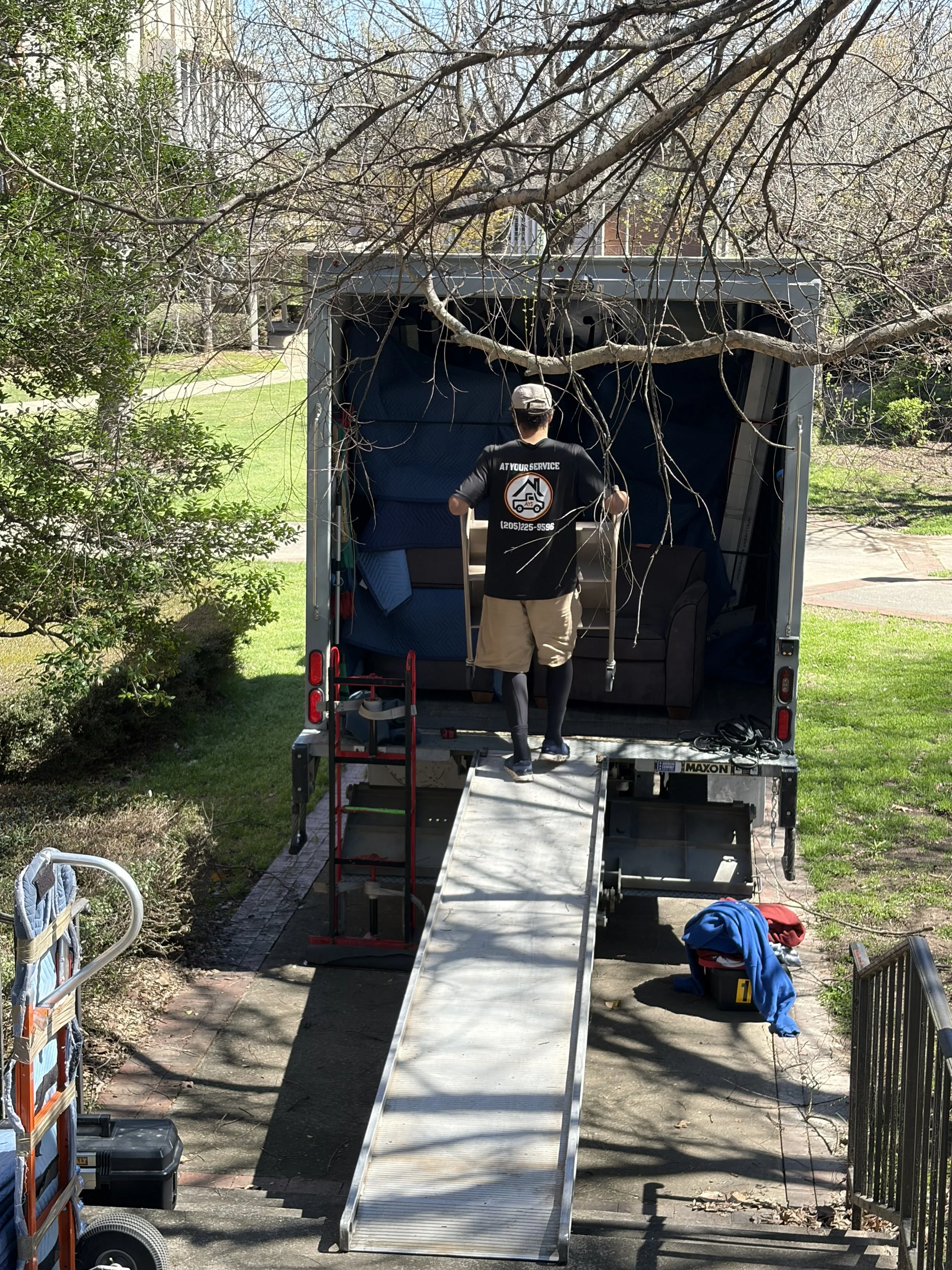 A mover loading furniture into a moving truck parked in a residential area with greenery, including trees and bushes, on a sunny day.