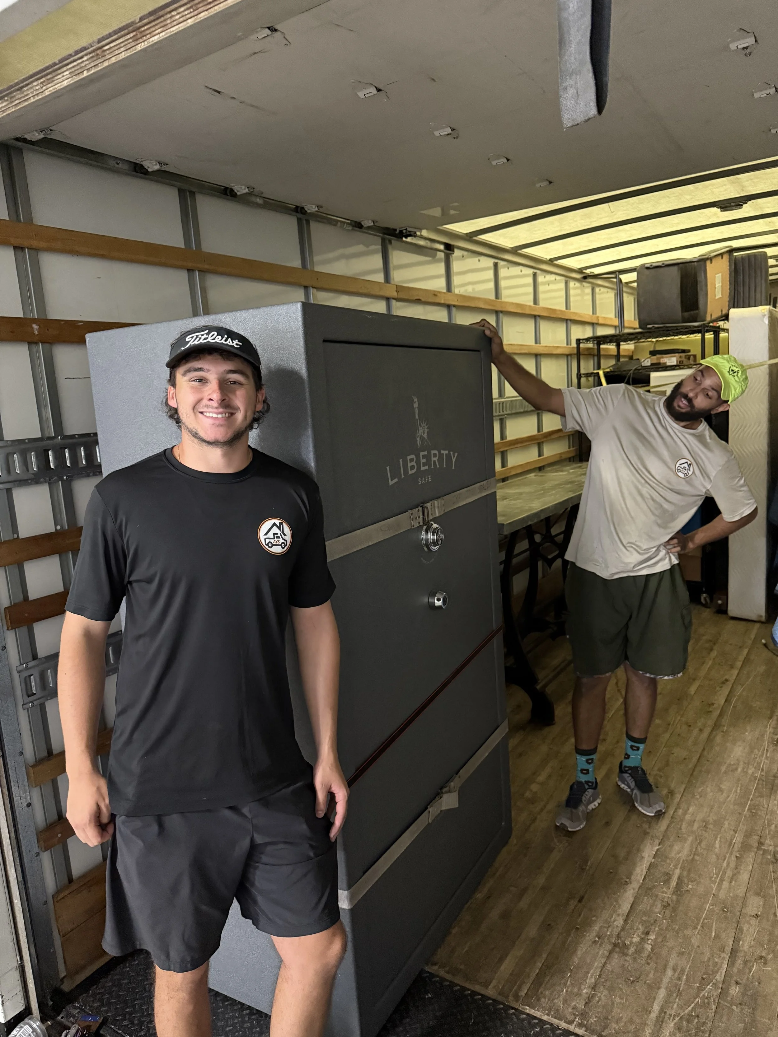 Two young men standing next to a large gray Liberty safe inside a moving truck. One is smiling at the camera, wearing a black T-shirt and shorts, and the other is leaning on the safe, wearing a beige T-shirt, shorts, and a bright green hat.
