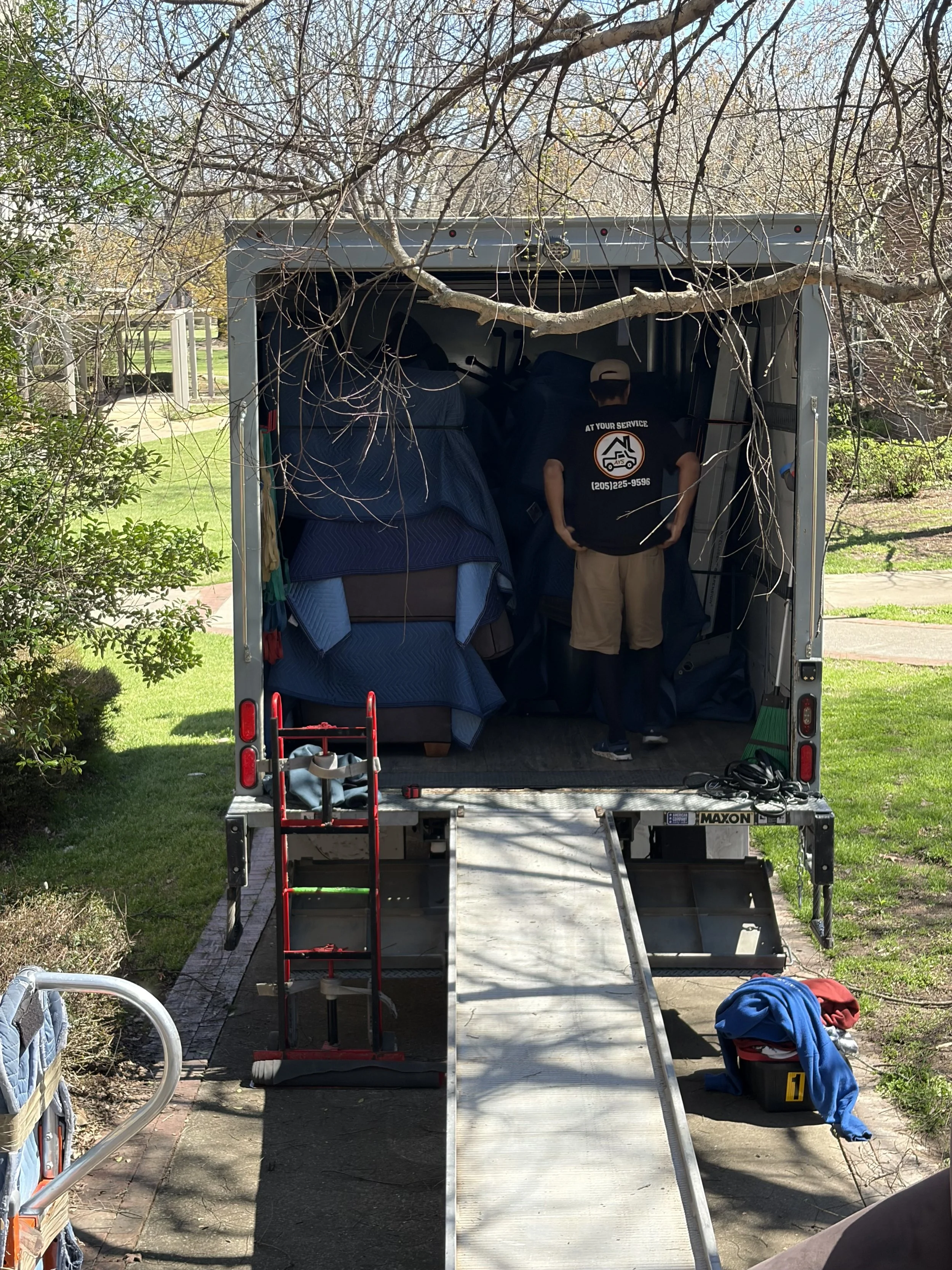 A moving truck parked on a driveway with the back open. Items like furniture and a mattress are loaded inside. A person wearing a black shirt, beige shorts, and a cap stands inside the truck with their back to the camera.