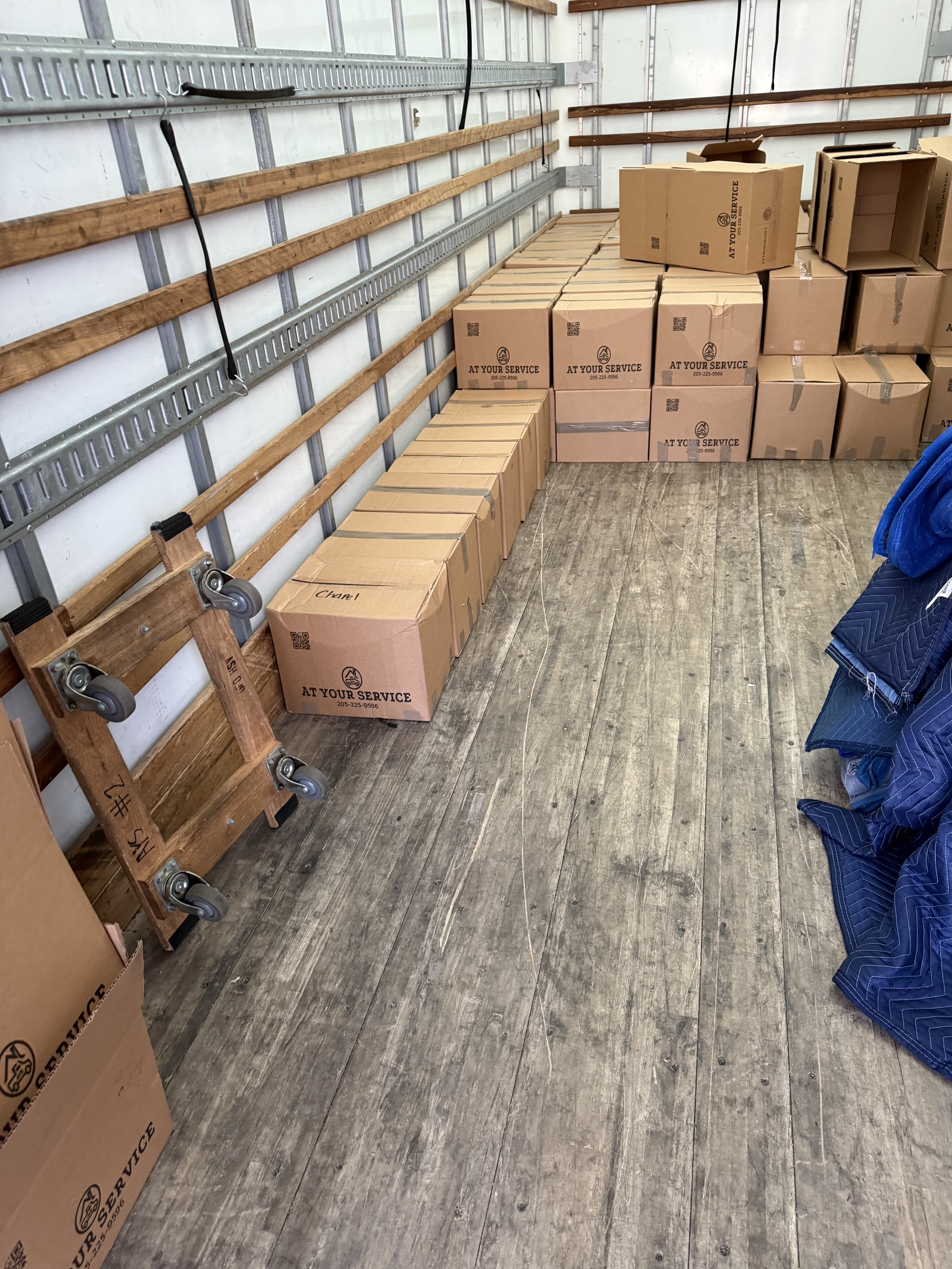 Empty truck interior with cardboard boxes stacked against the walls, some labeled 'AT YOUR SERVICE'. A folded dolly and a blue blanket or tarp are also visible.