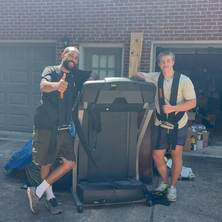 Two young men stand outside a garage, leaning on a treadmill, smiling and giving thumbs up.