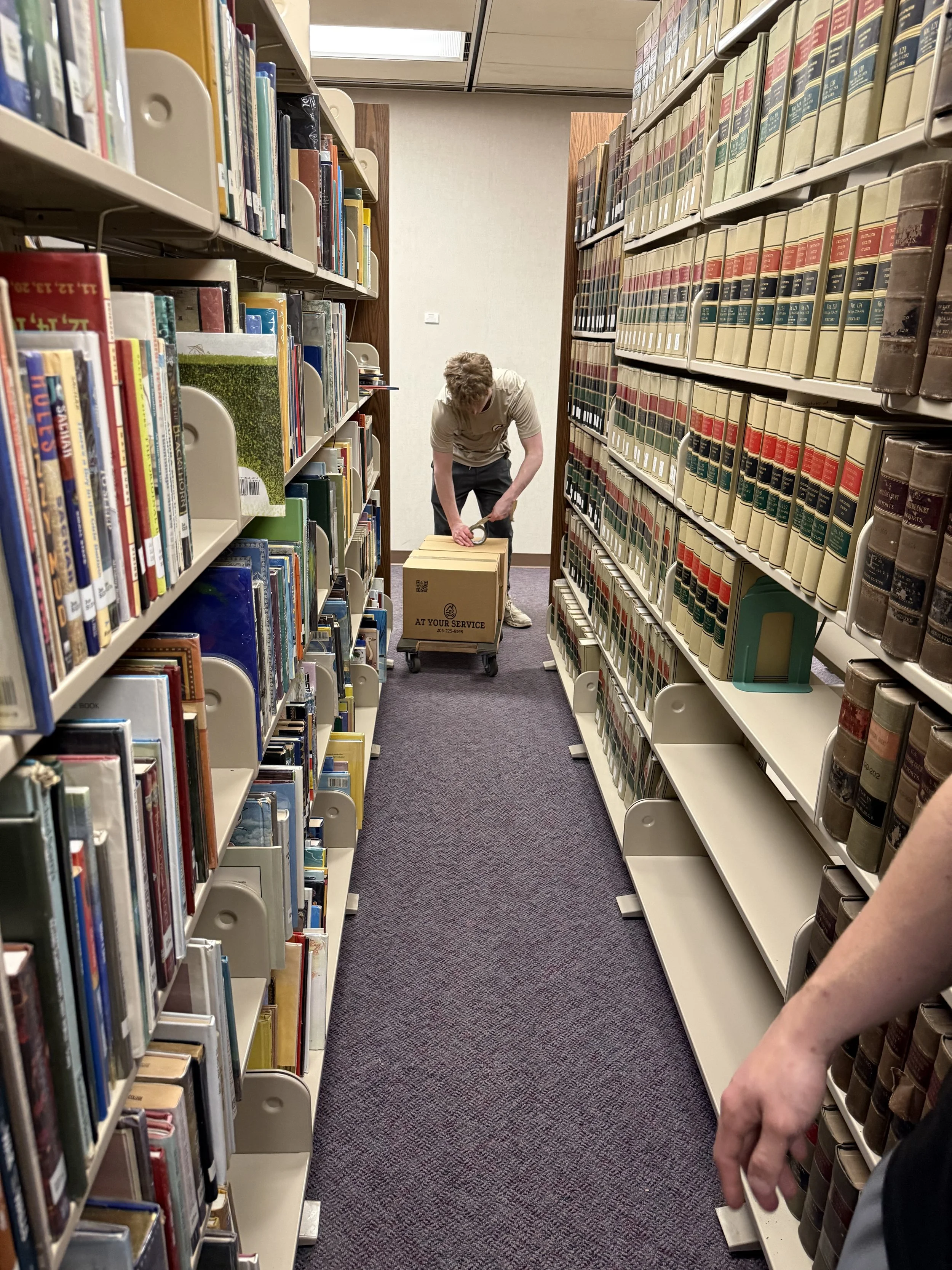 A person using a hand truck to move a box in a library aisle, surrounded by shelves filled with books.