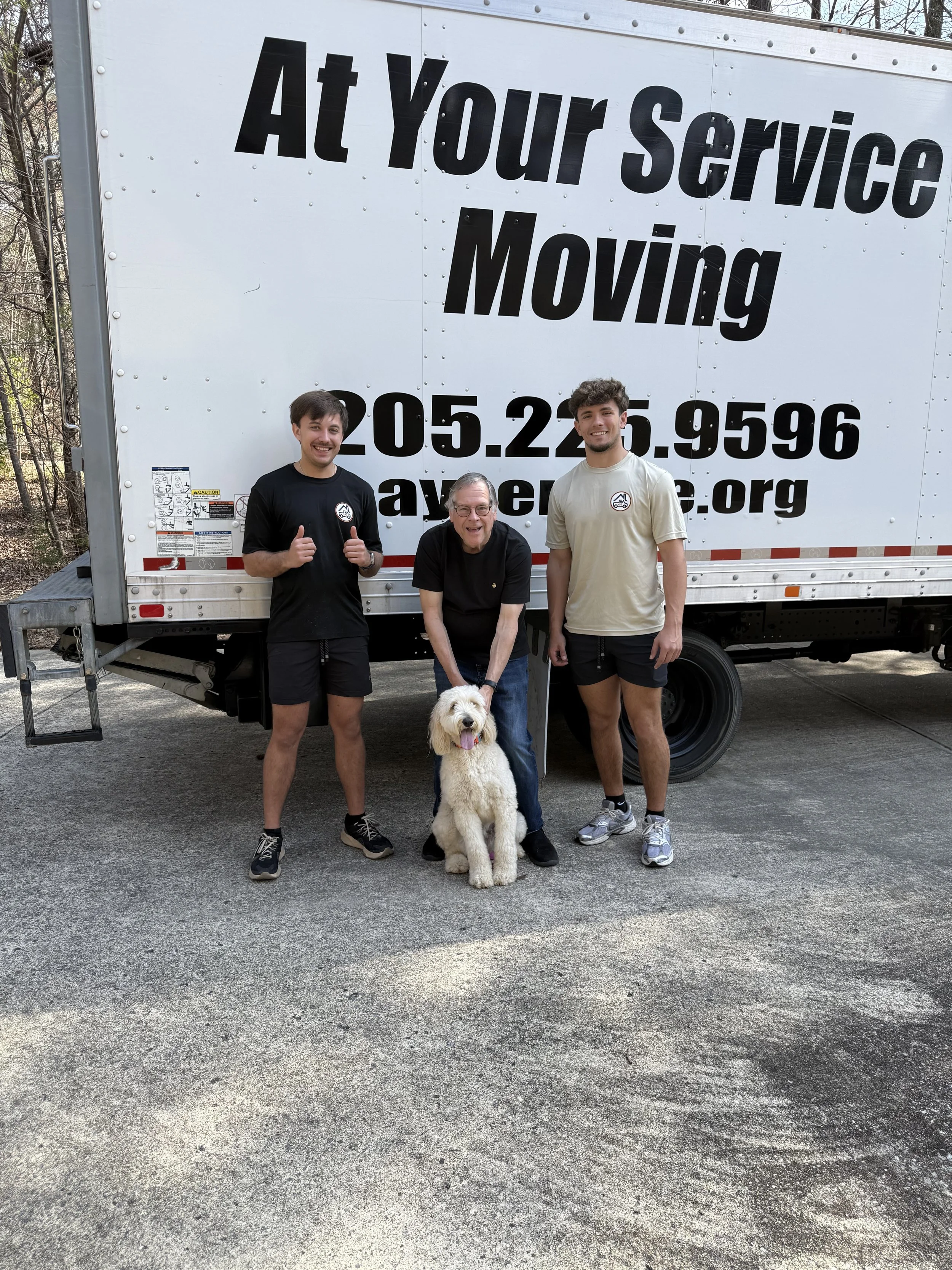 Three men and a dog standing in front of a moving truck with a large sign. The men are smiling, with the two on the sides giving thumbs up. The sign on the truck reads "At Your Service Moving" along with a phone number and website.