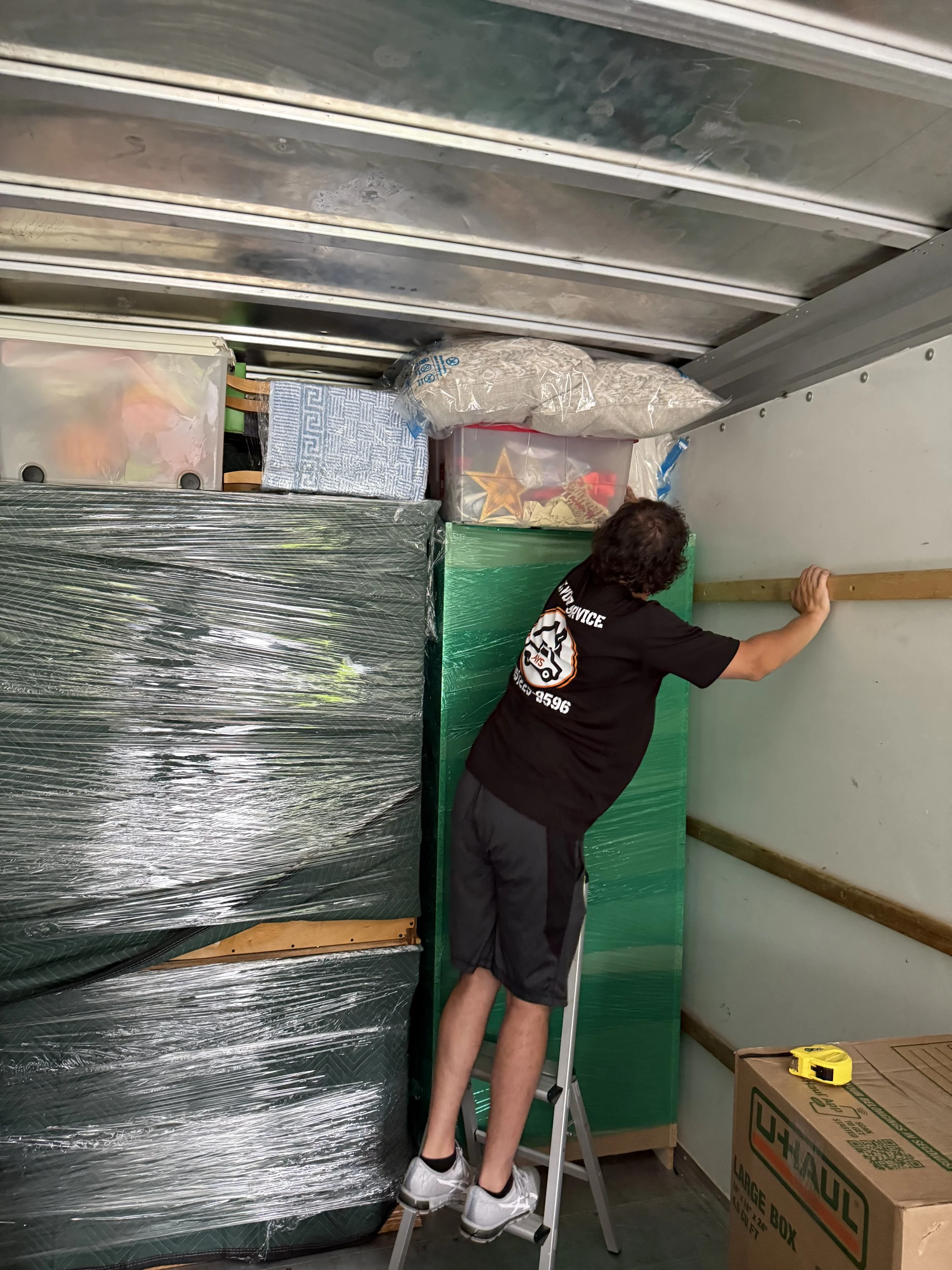 Person on a ladder measuring a wall inside a storage unit filled with wrapped furniture, boxes, and miscellaneous items.