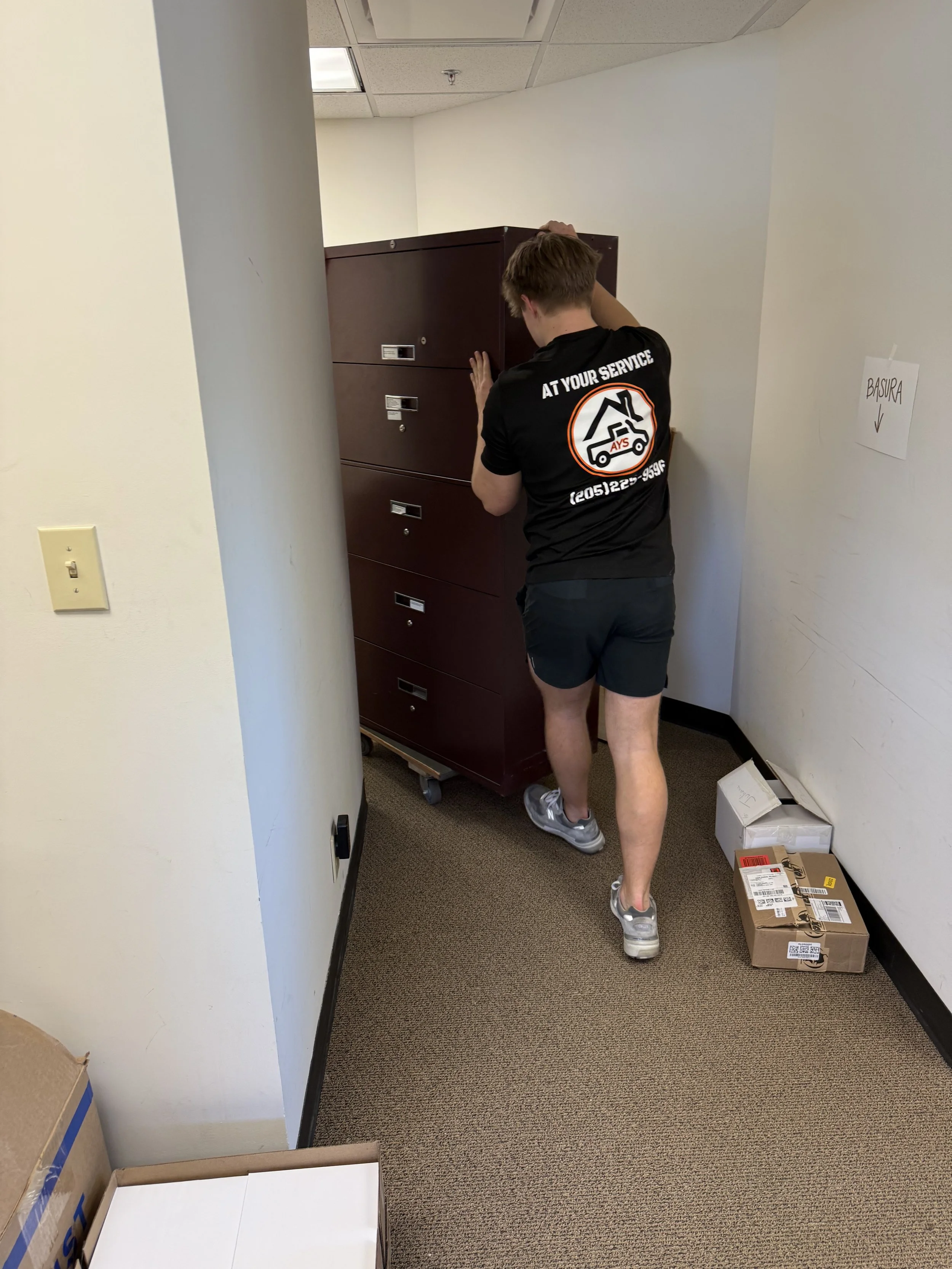 A mover wearing a black t-shirt and shorts pushes a brown filing cabinet on wheels through an office hallway. The back of the shirt has a logo and text that reads 'At Your Service' and a phone number.