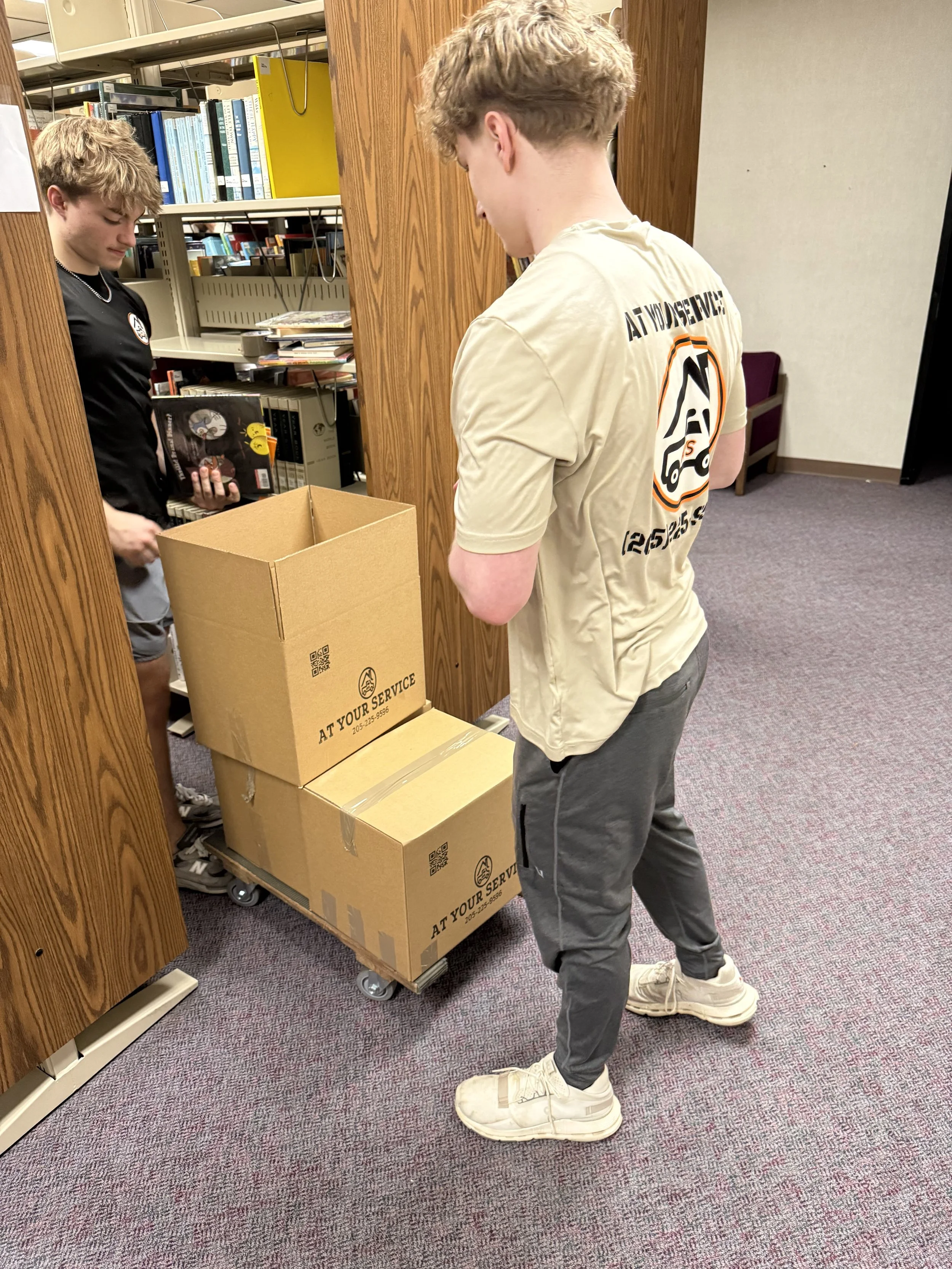 Two young males in a library or office setting, one holding a library book and the other standing beside a dolly with cardboard boxes, one open, labeled "AT YOUR SERVICE."