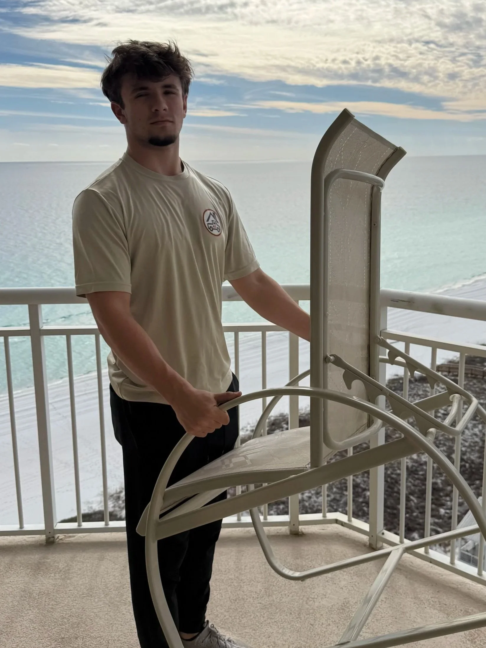 A young man with dark, wavy hair stands on a balcony by the ocean, holding a beige lounge chair with a built-in canopy.