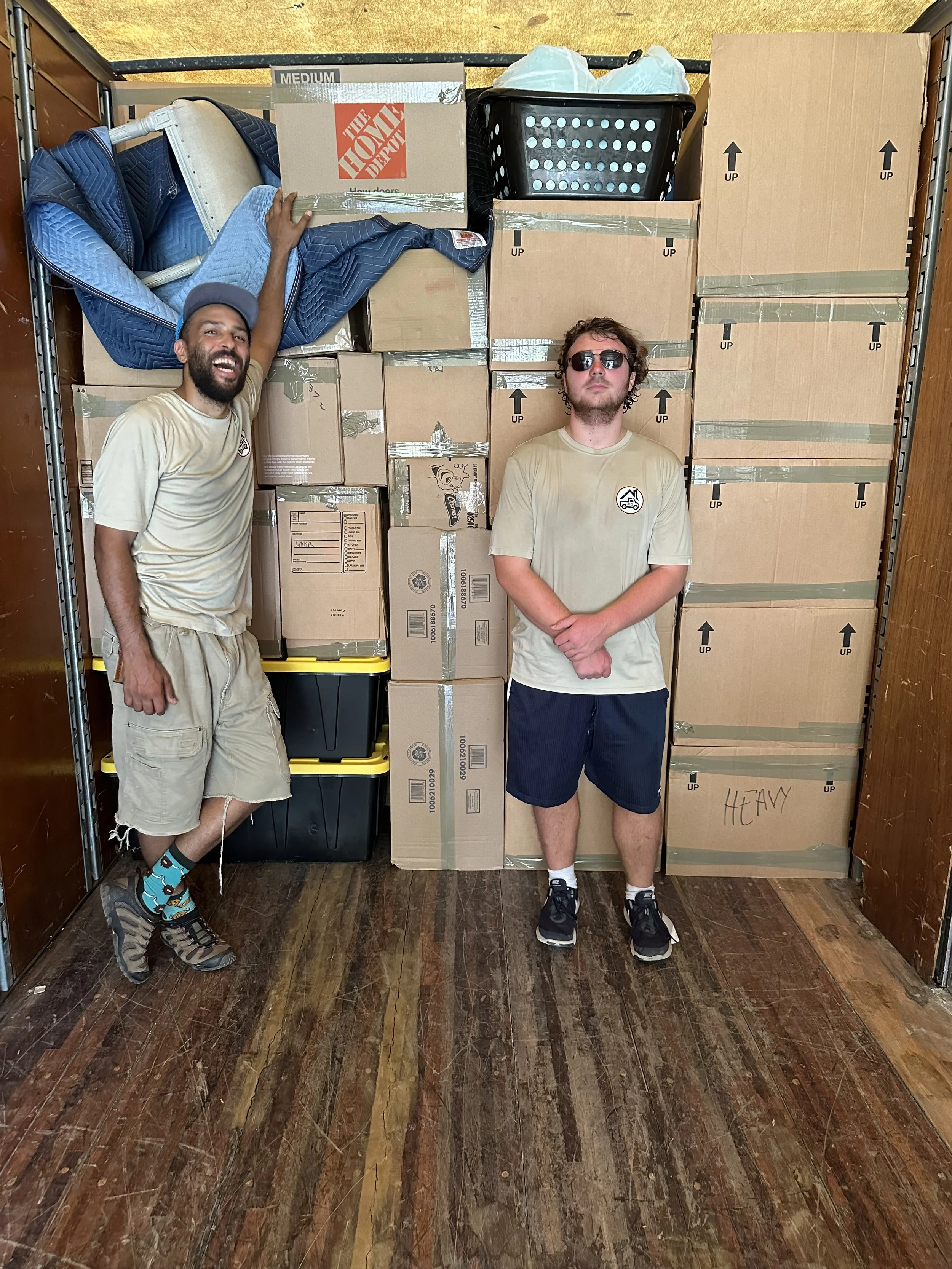 Two young men are posing in front of a large stack of boxes and storage containers. One is smiling and reaching up, while the other stands with arms crossed, wearing sunglasses.