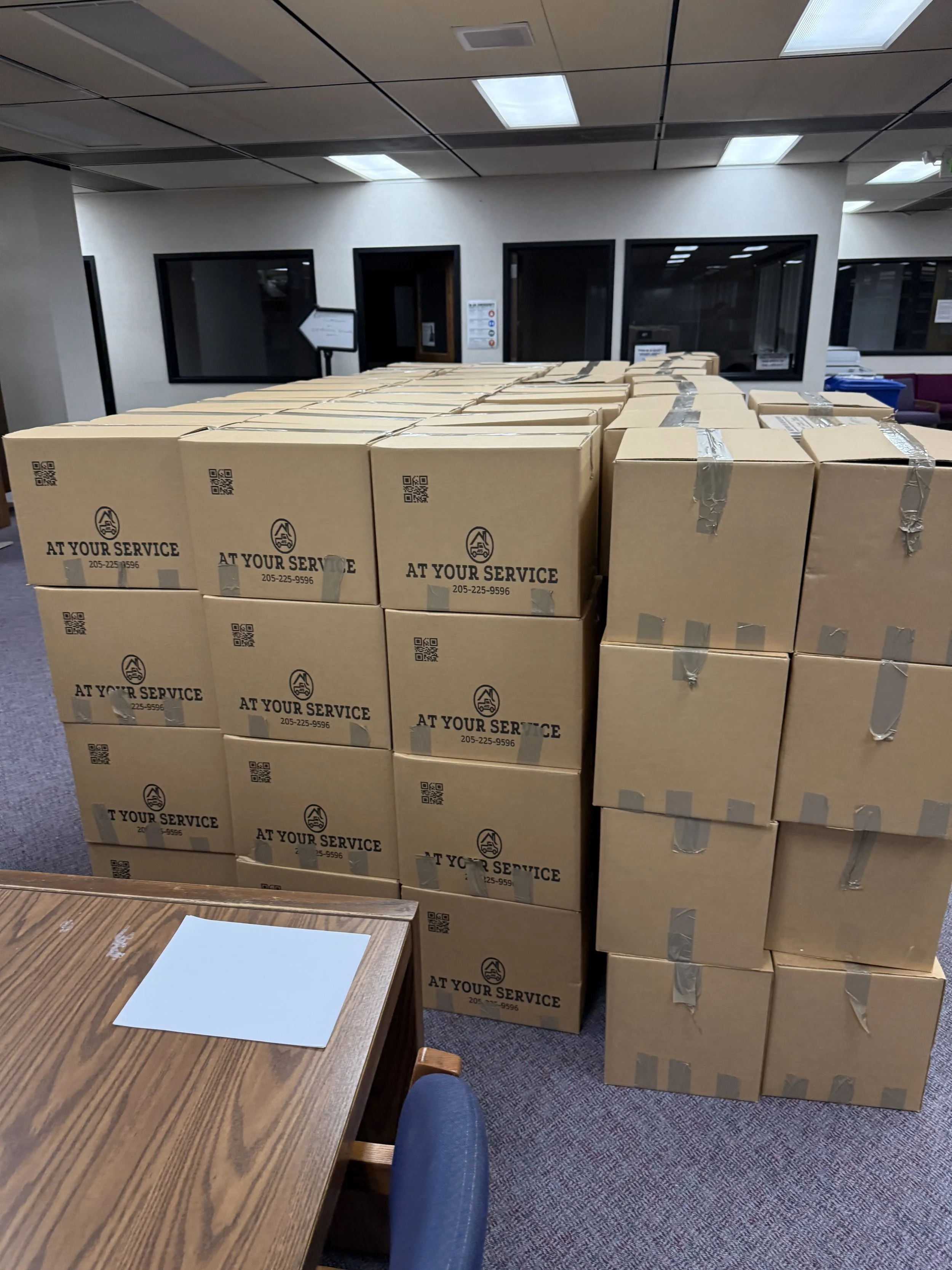 Stack of cardboard boxes in a room, labeled with 'AT YOUR SERVICE' and contact number, with a wooden table and chair in the foreground.