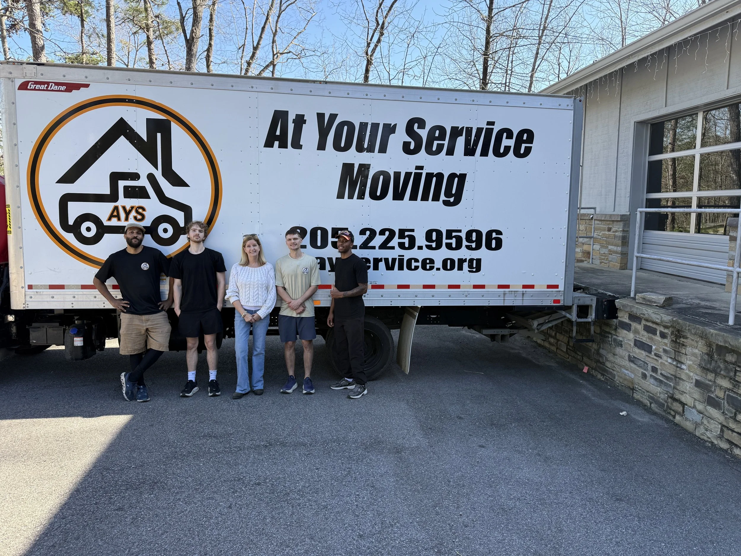 A group of five people standing in front of a moving service truck, outdoors on a driveway, with trees and a building in the background.