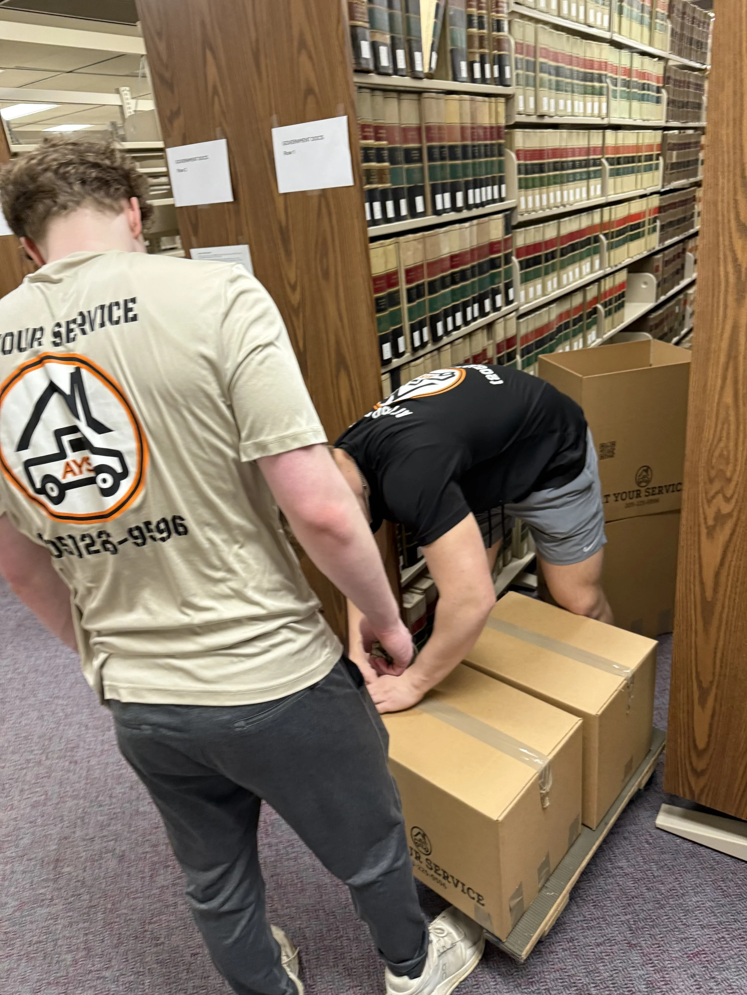 Two delivery workers are packing boxes in a library storage area. One person is standing with their back to the camera, wearing a beige t-shirt with a logo and text, while the other is bent over, sealing boxes on a cart, with shelves of books in the 