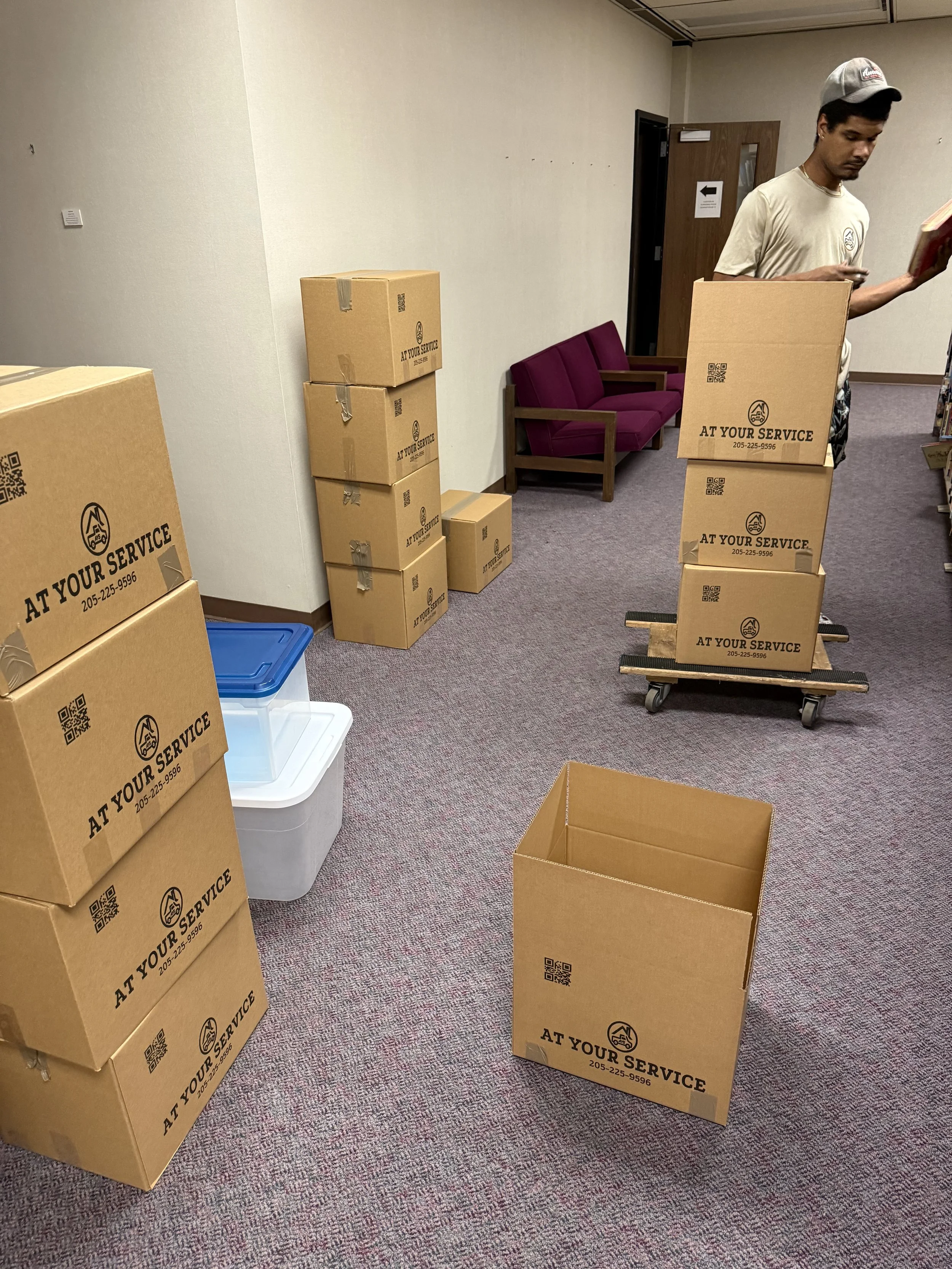 A man with a cap looking at his phone, standing next to stacked cardboard boxes labeled "At Your Service" in a room with purple chairs and storage bins.