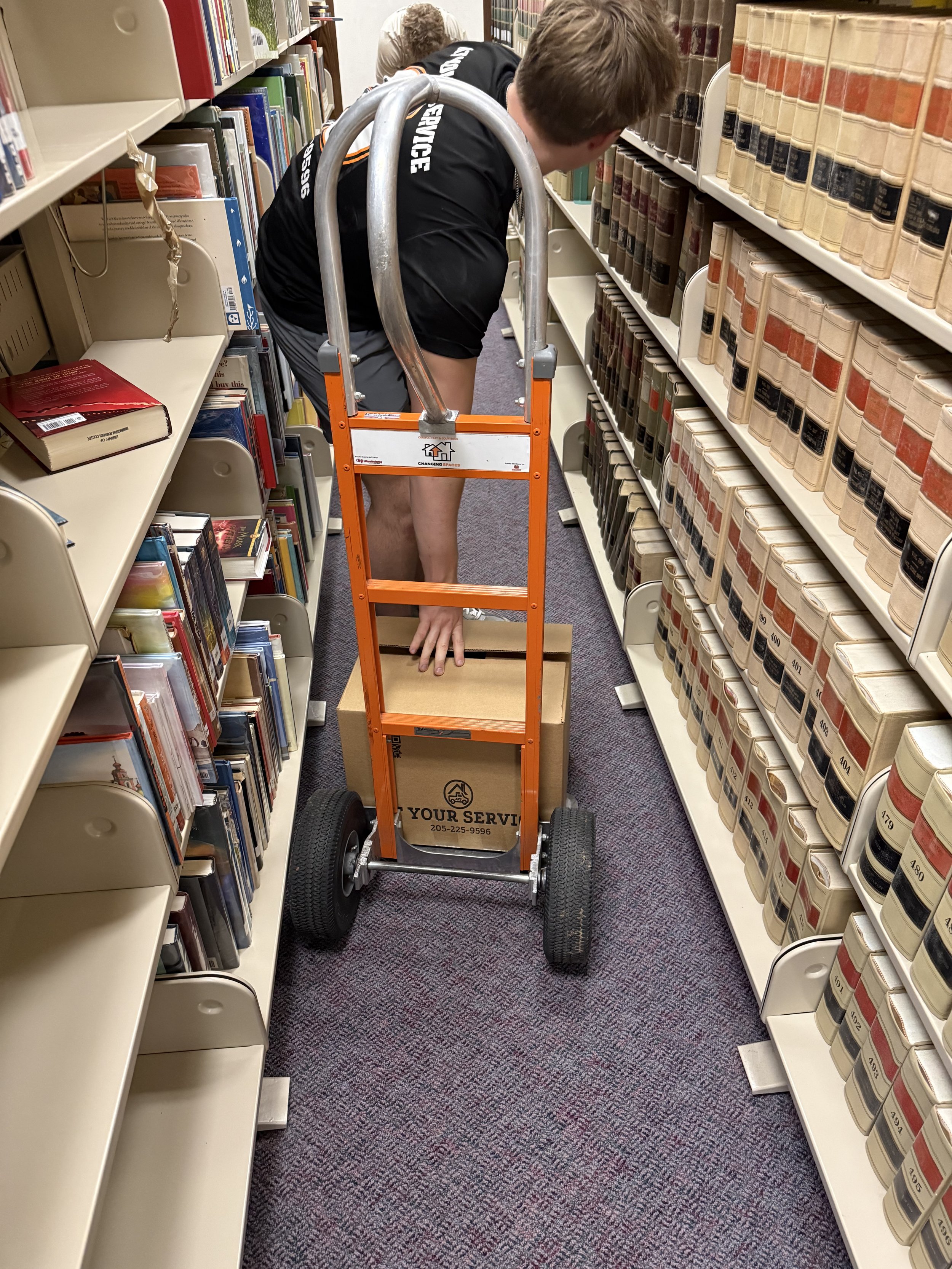 A person in a library is using a hand truck to move a cardboard box between bookshelves. The person is bending over and reaching into the box. The shelves are filled with books, some of which are labeled.