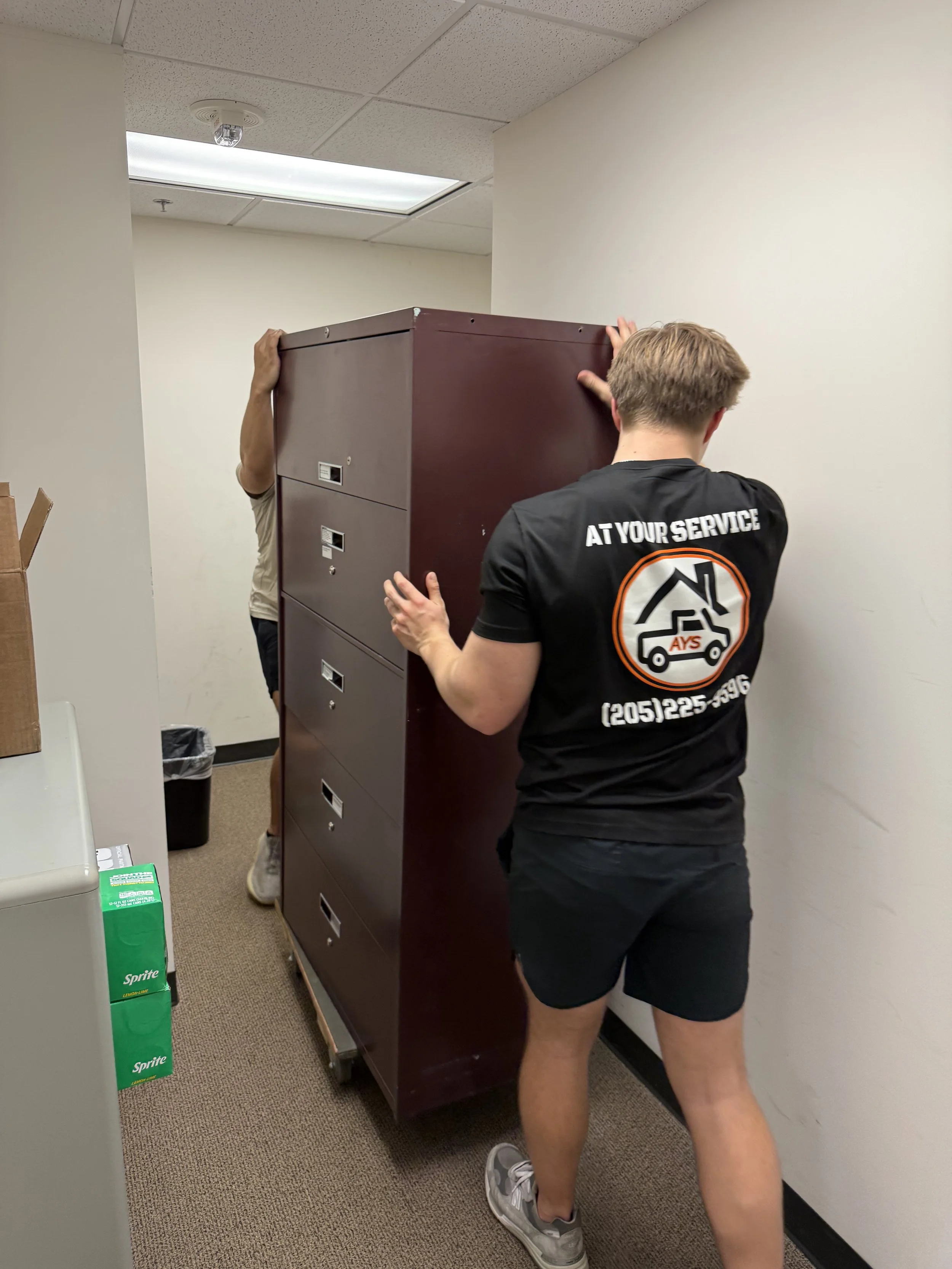 Two people are moving a large, dark red filing cabinet in an office hallway. One person is wearing a black shirt with a logo and phone number on the back, and the other is partially hidden behind the cabinet.