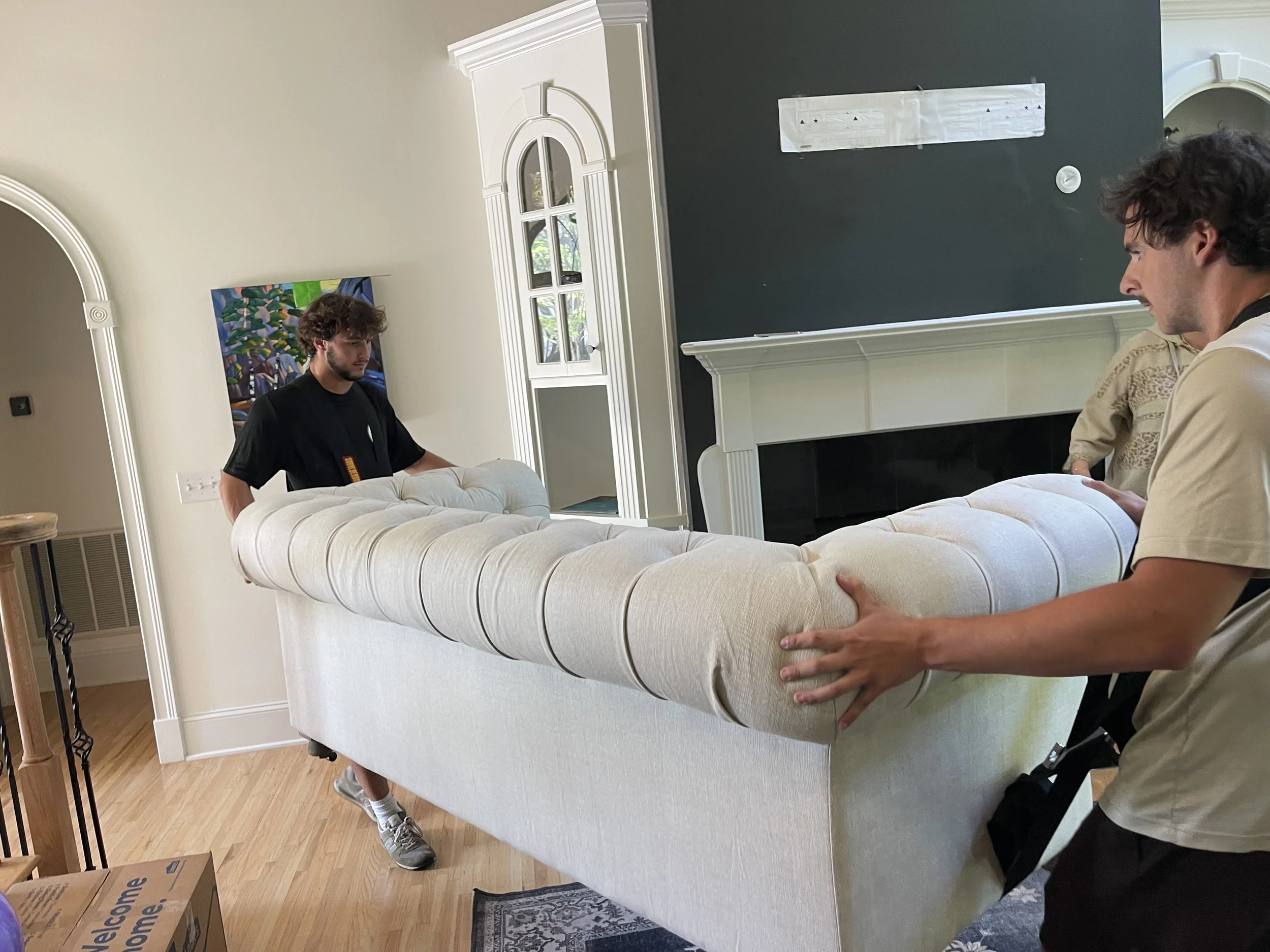 Three young men are moving a large, light-colored couch into a living room indoors. One man is on the right side, holding the arm of the couch, while another man is on the left side, gripping the other arm. The third person is partially visible behin