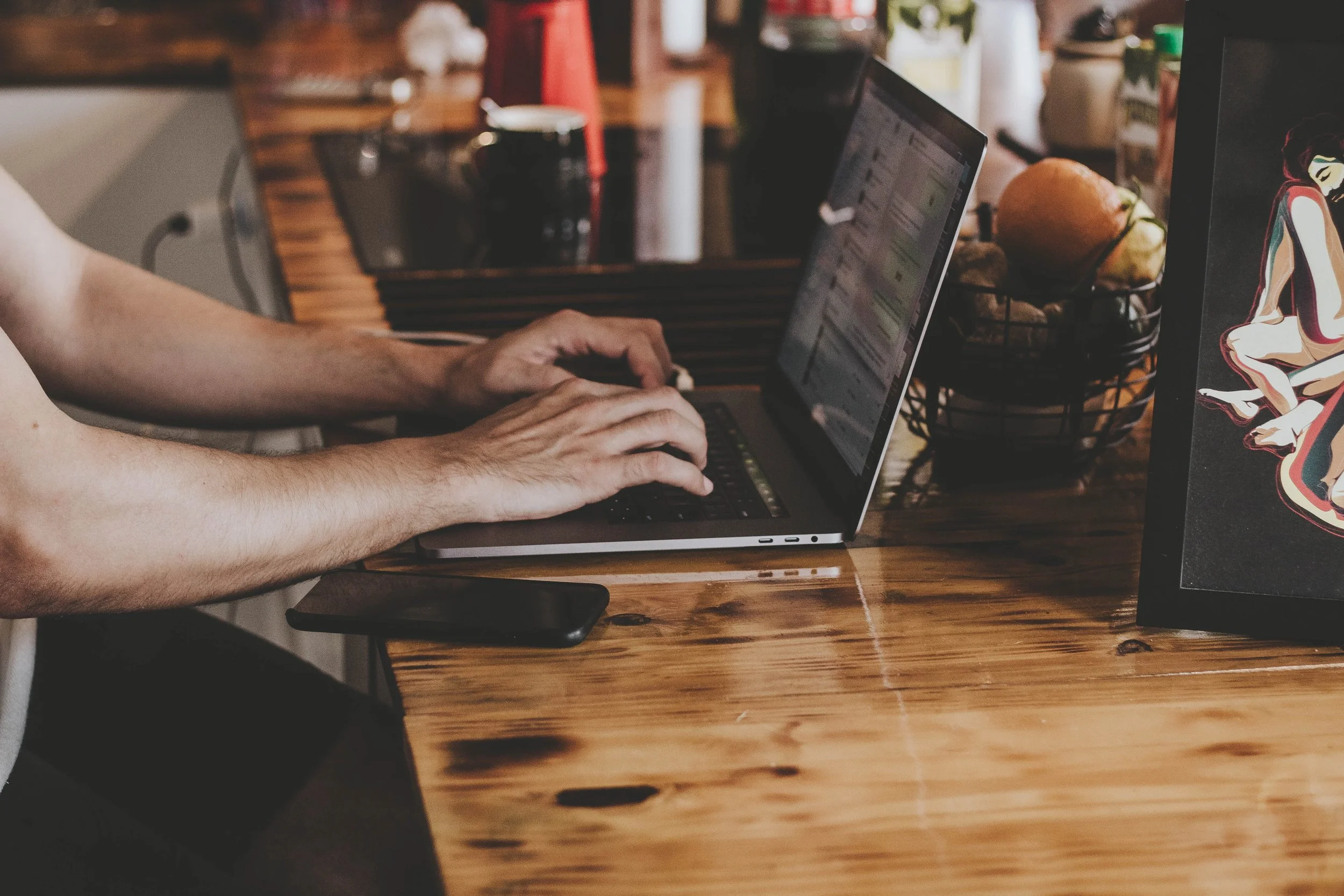 Person typing on a laptop at a wooden table with a fruit basket and framed artwork.