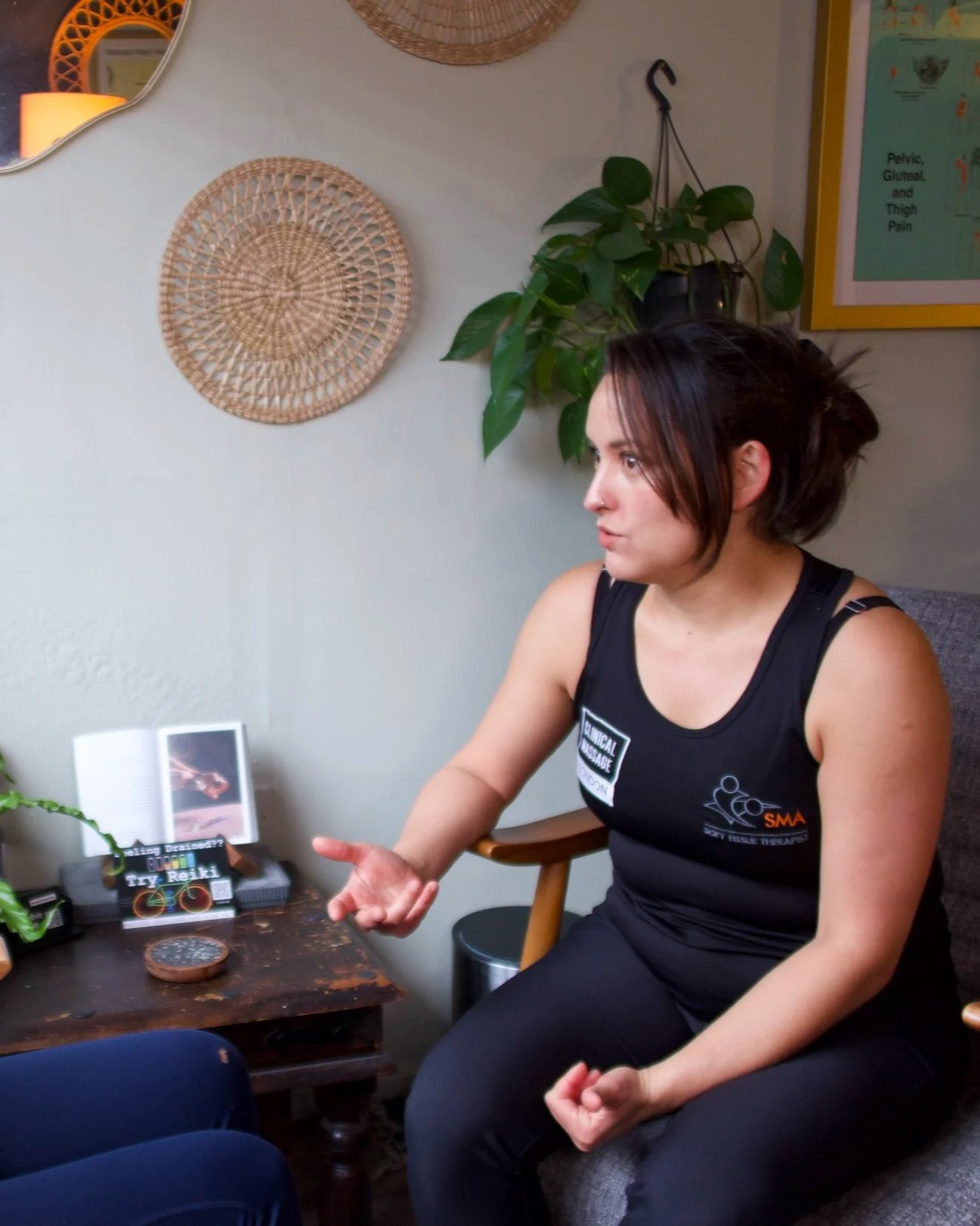 A woman sitting on a couch, gesturing with her left hand, engaged in conversation, with plants, wall art, and a small wooden table with a book and digital device nearby.