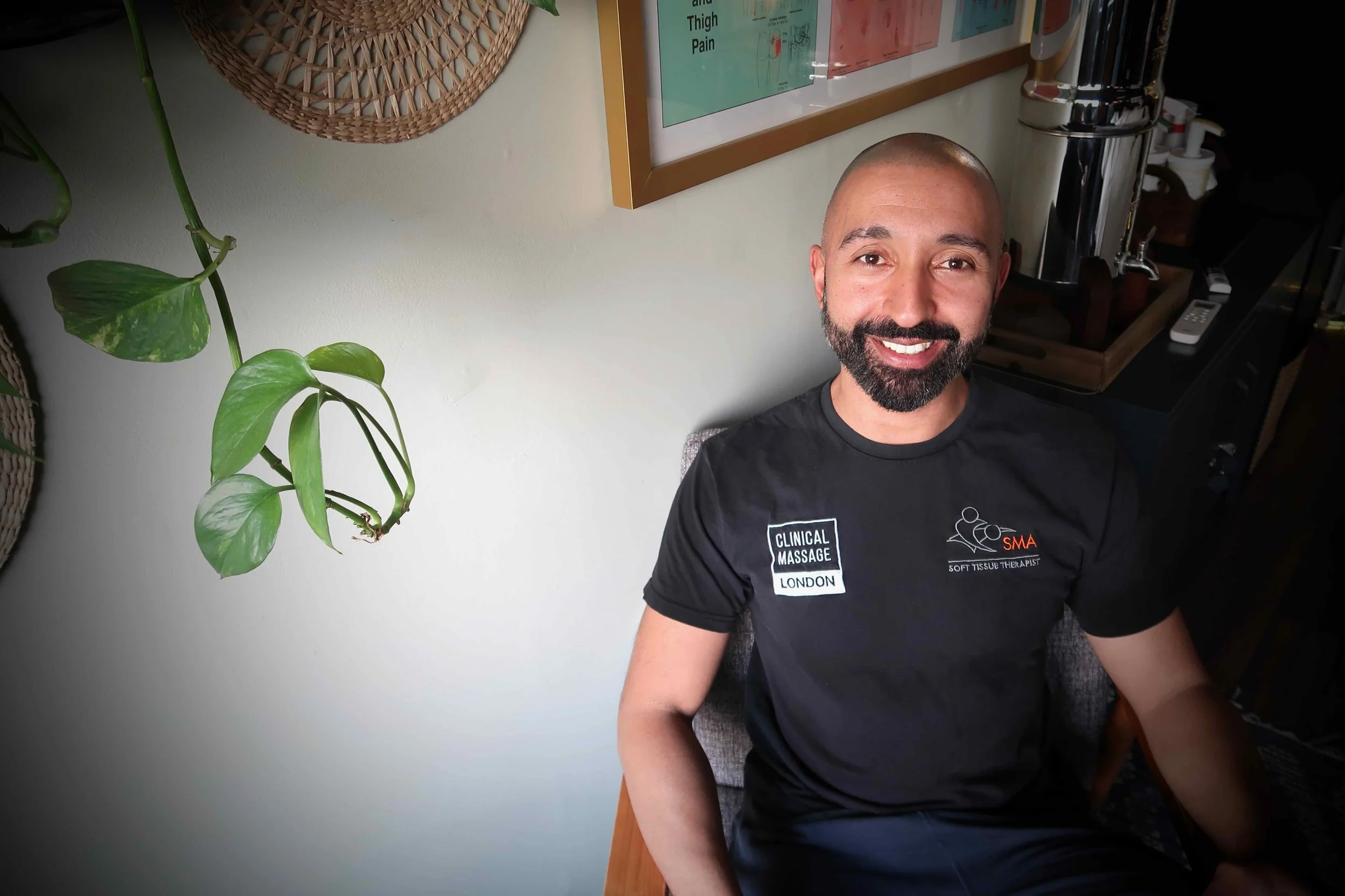 Sports Massage therapist Steve Berry  sitting indoors near a light-colored wall with plant and framed chart, wearing a black shirt with logos for clinical massage London and soft tissue therapy.