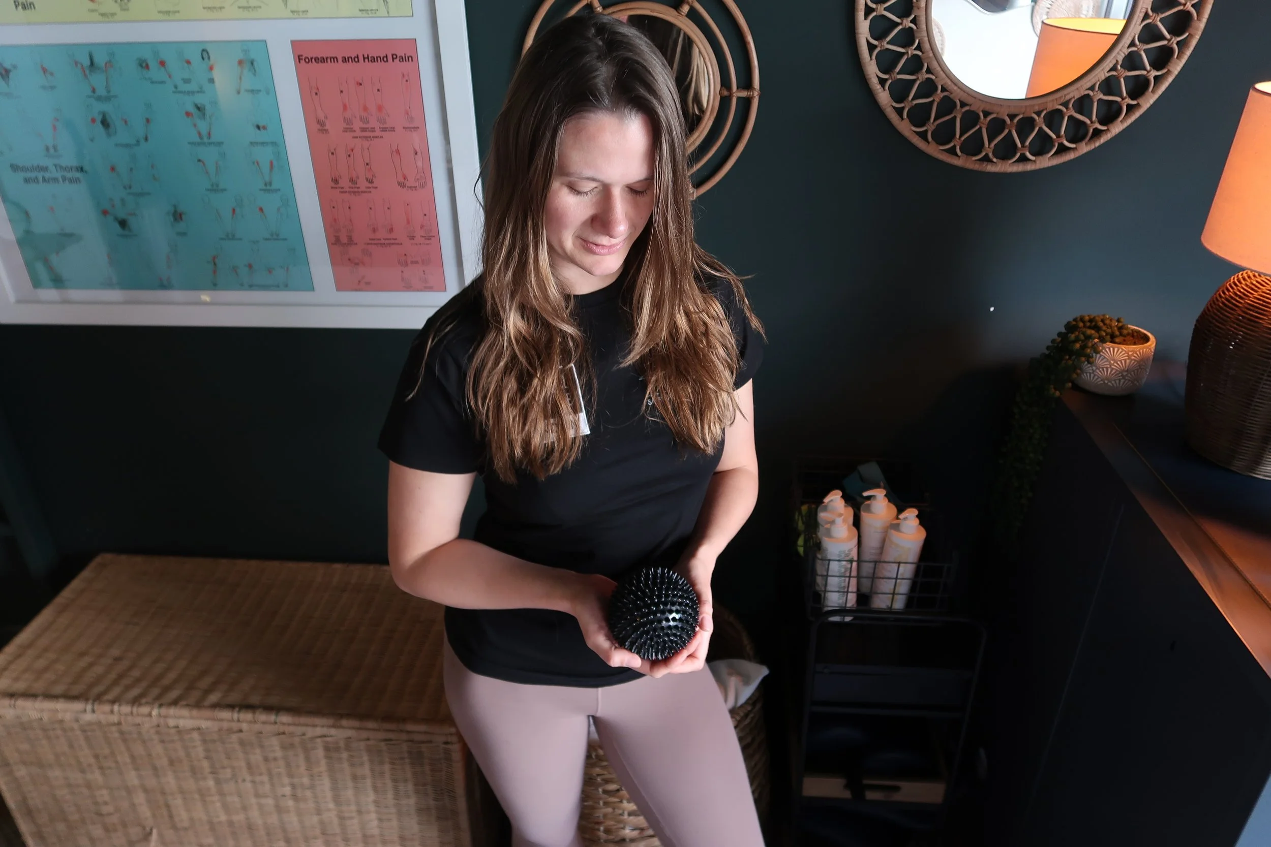 A woman in a black shirt holding a black textured massage ball in a room with framed mirrors, a wicker storage bench, and a dark wall with a medical chart.