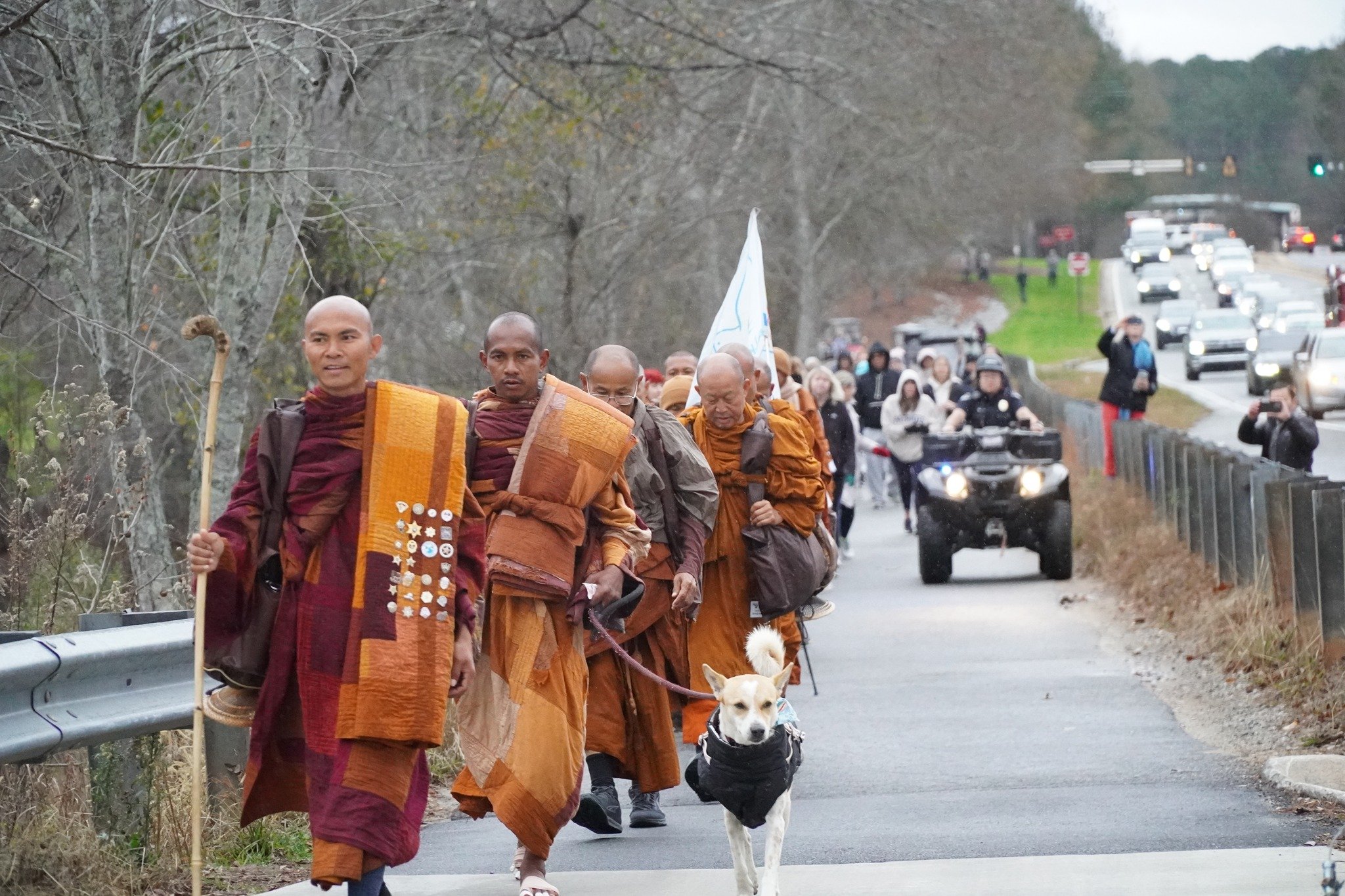 A group of Buddhist monks in orange and maroon robes walk along a roadside during a peace march, led by one holding a staff, with a small dog in a jacket trotting beside them. A crowd and vehicles follow behind on the road.