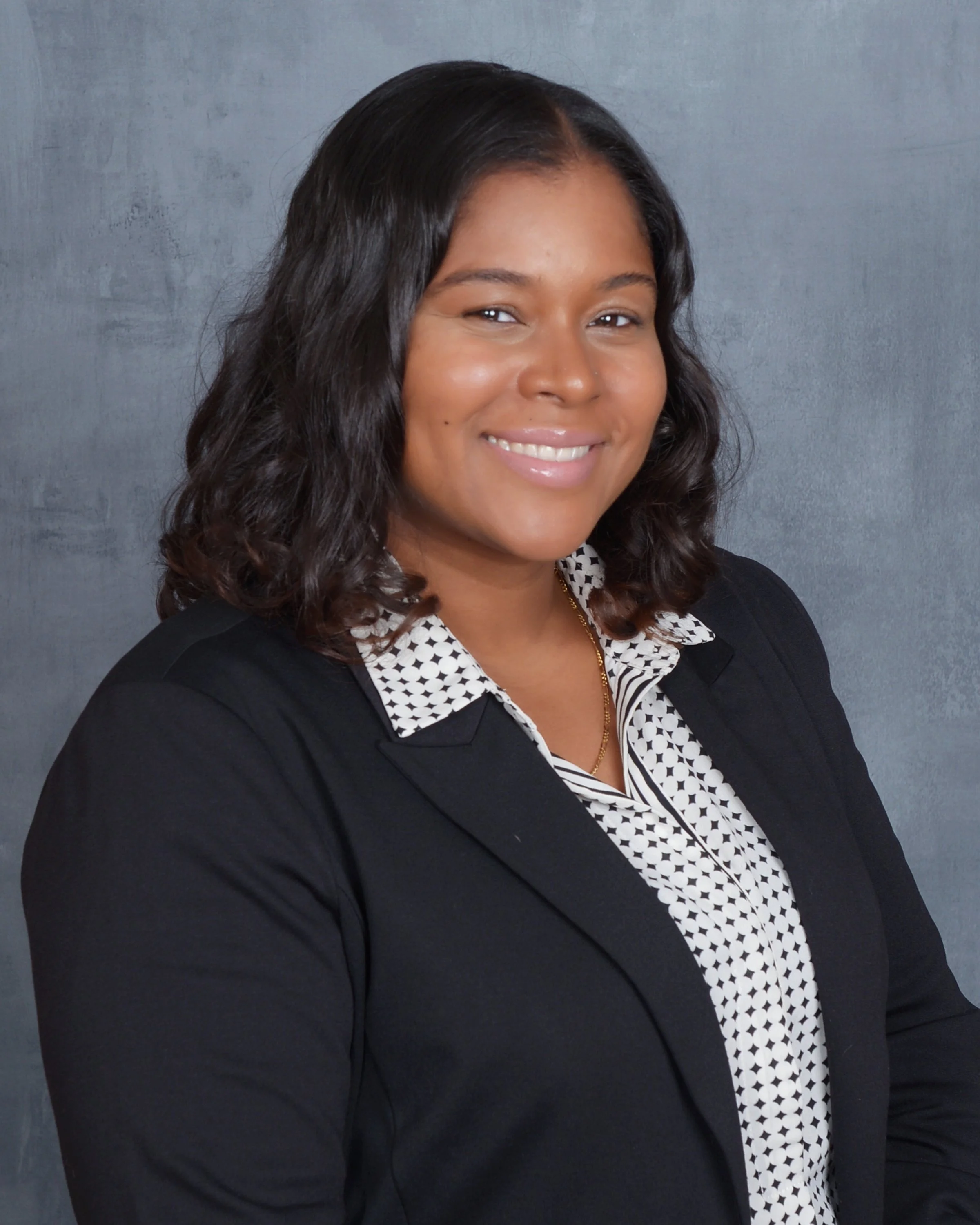 A woman with medium skin tone and shoulder-length black hair, wearing a black blazer over a white blouse with black polka dots, smiling against a gray textured background.
