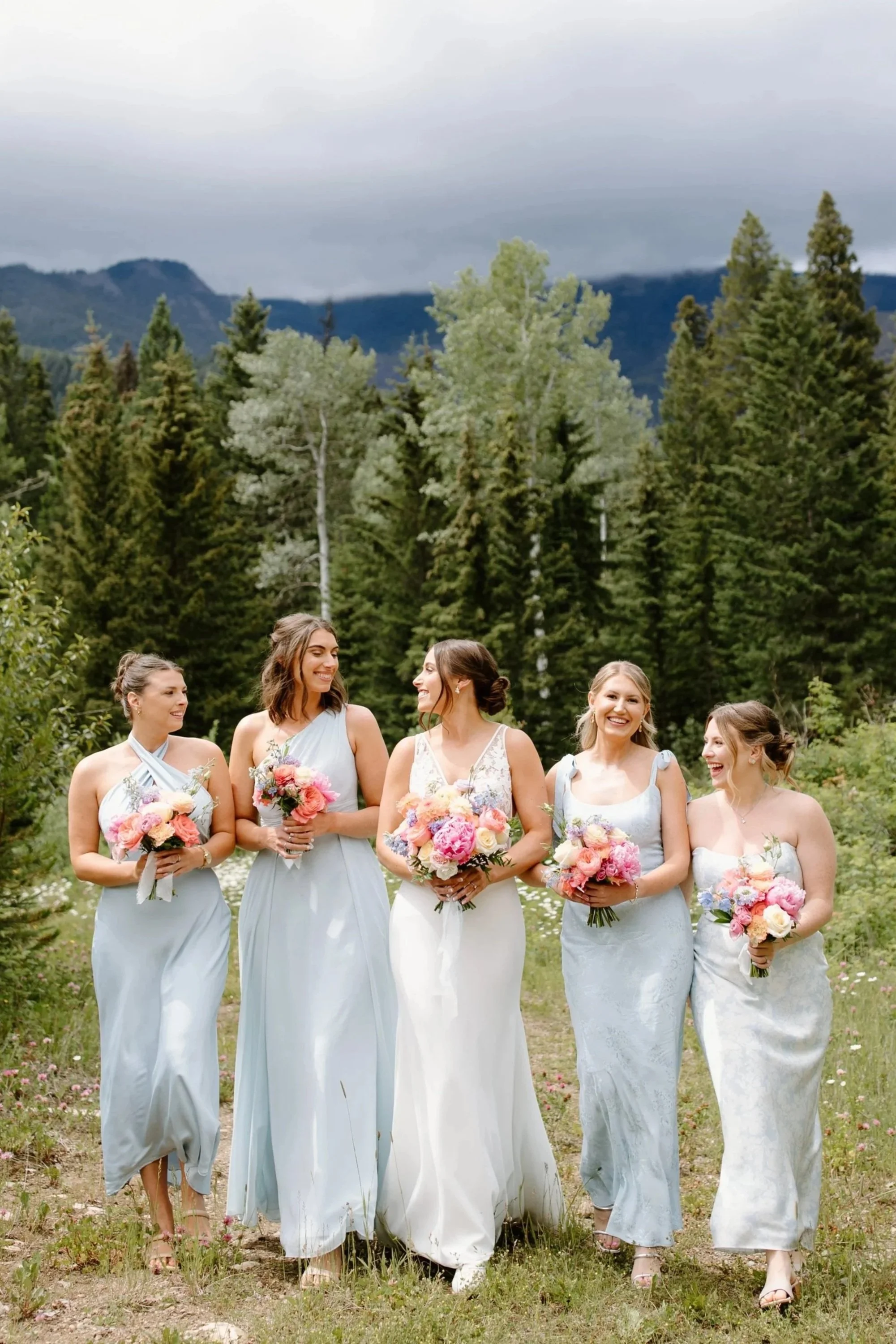Bride and bridesmaids walking through a meadow holding bright bouquets, wearing soft blue dresses at bride's western chic barn wedding