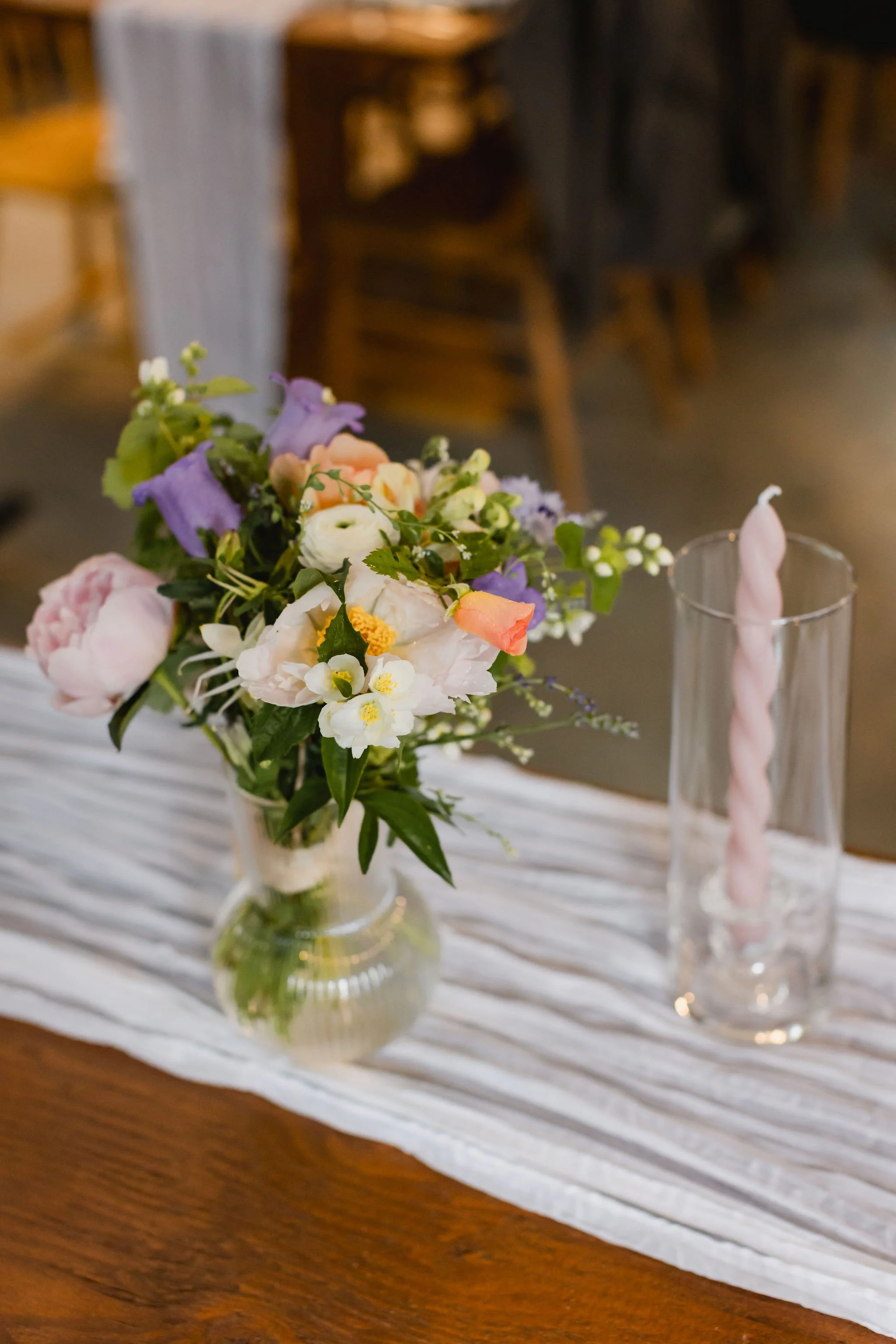 Pastel floral centerpiece with lilac, peach, and white blooms beside a blush taper candle at a Western Chic BC wedding.