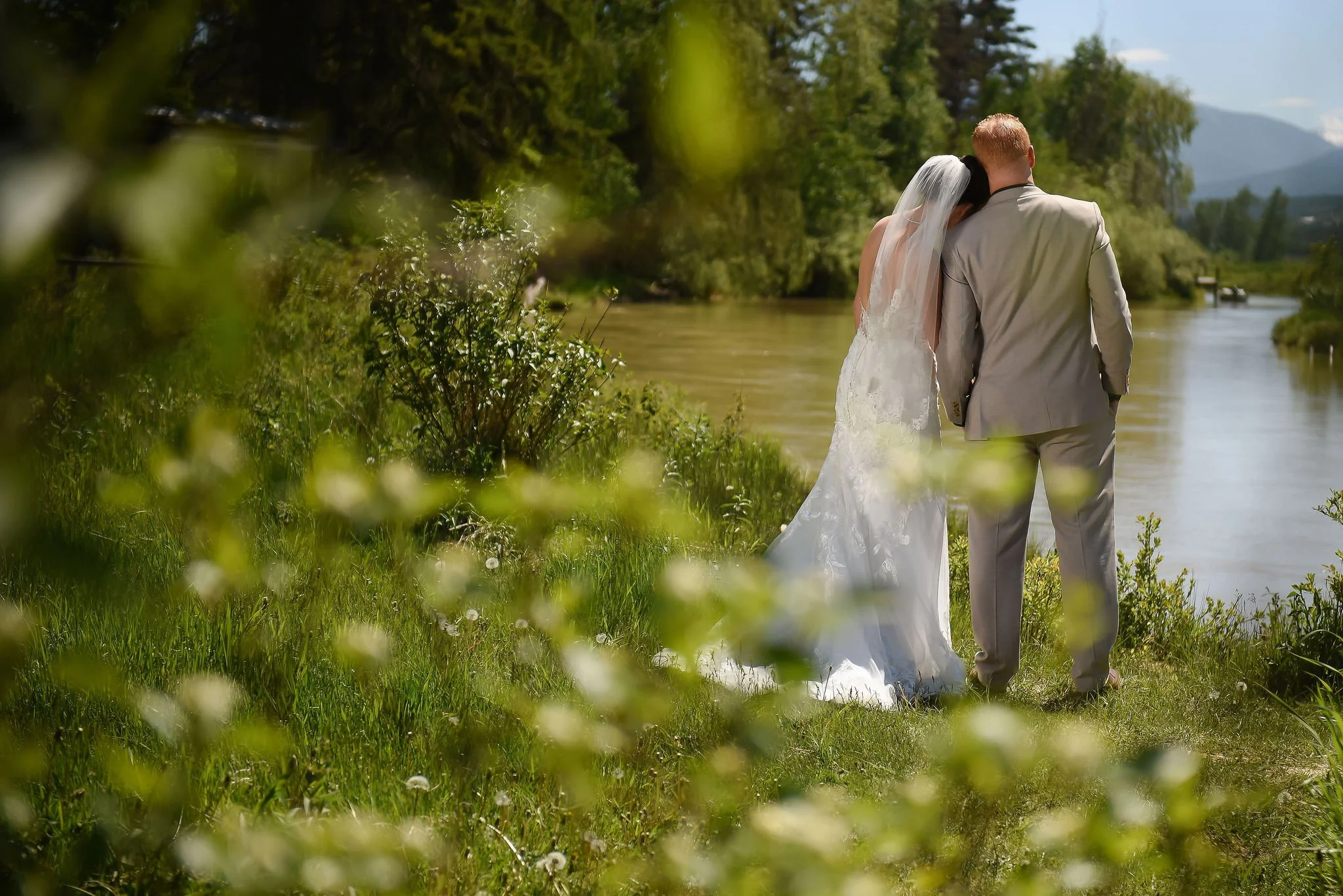 The bride leans on the groom's shoulder as they face the river during a quiet moment at their ski resort wedding