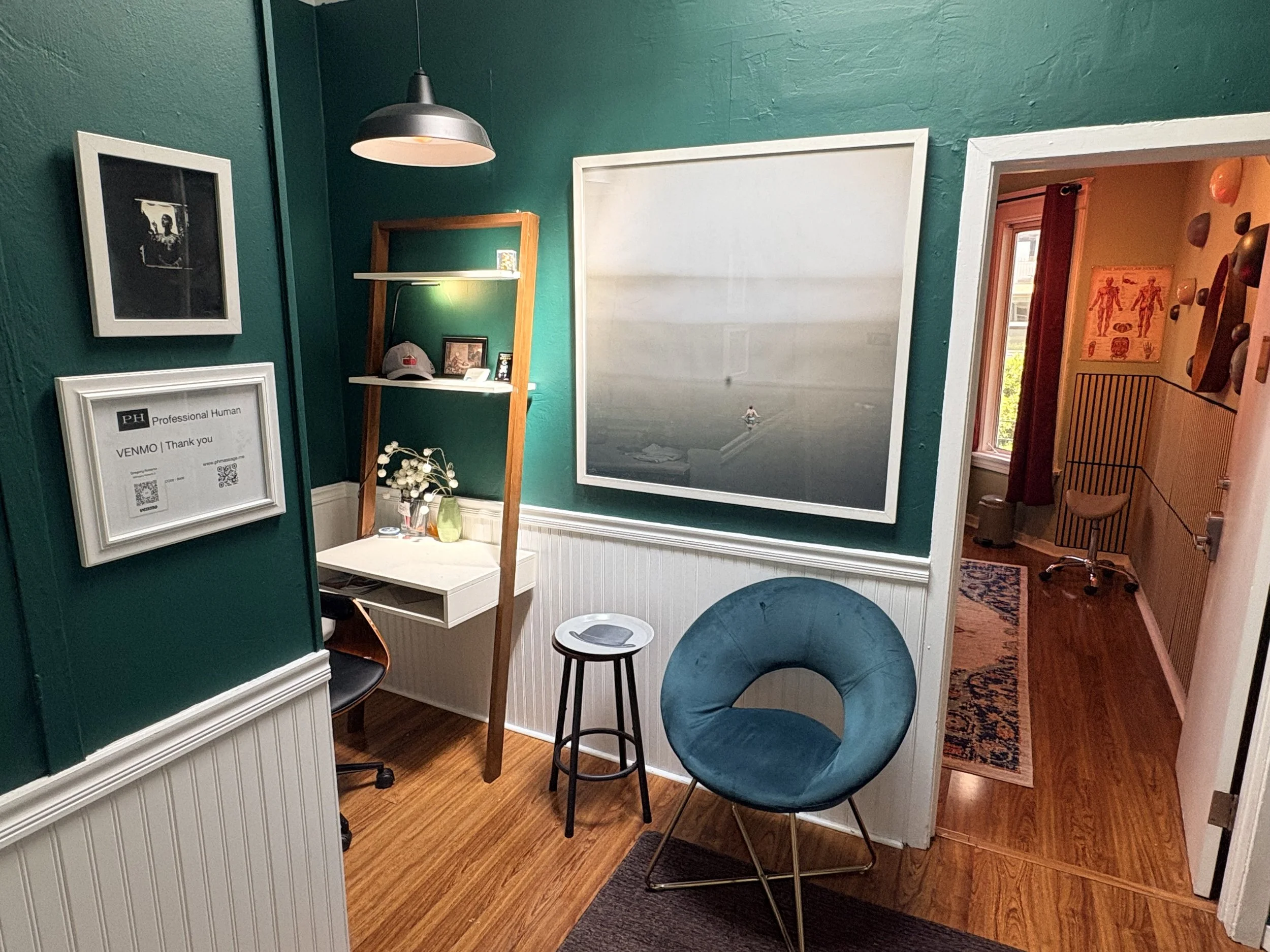 Check out lobby, 3rd floor, Interior view of a cozy room with teal walls, a large framed mountain landscape photo, a small white desk with a chair, a blue round chair, shelves with decor items, and an adjacent room with medical posters and a window.