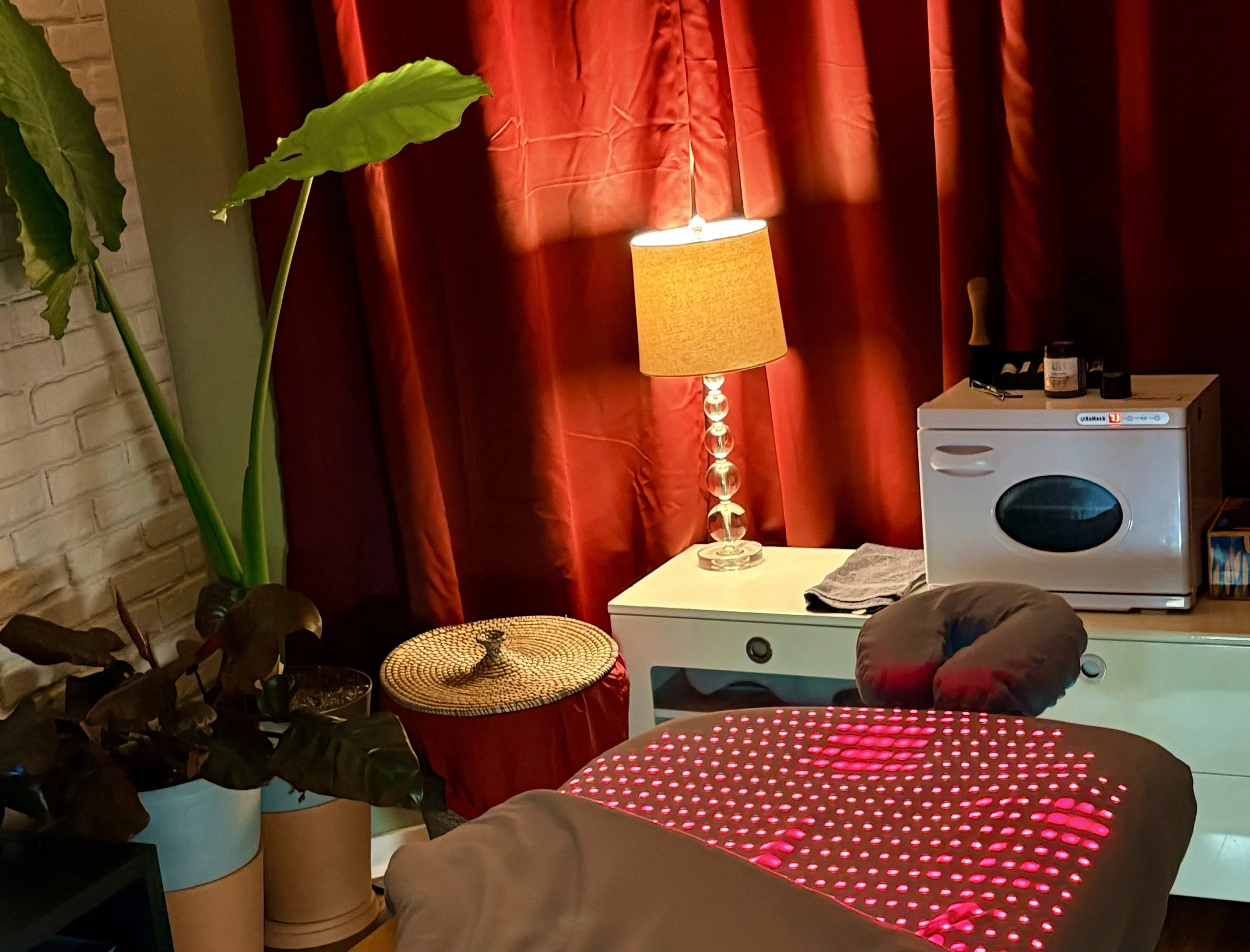 Interior of a room with red curtains, a tall clear glass lamp with a yellow shade, a large green-leafed plant, a white table, a washing machine, and various household items.