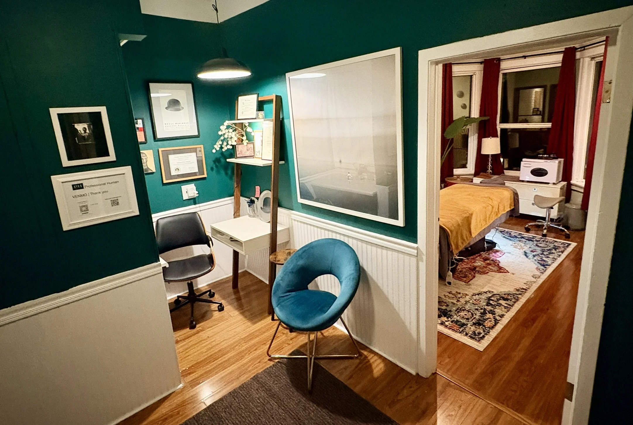 A cozy room with teal walls decorated with framed artwork, a small white desk, a black rolling chair, and a blue upholstered chair. There is a mirror and some shelves with knickknacks. The adjacent bedroom has a bed with a yellow blanket, red curtains, a small table with a lamp, and a window. A colorful rug covers part of the wooden floor.