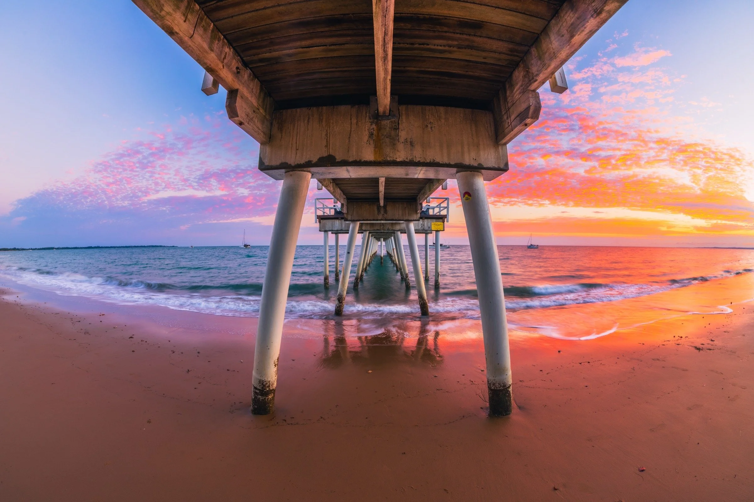 A view beneath Scarness Jetty as gentle waves meet the sand and pink and orange clouds light the evening sky.