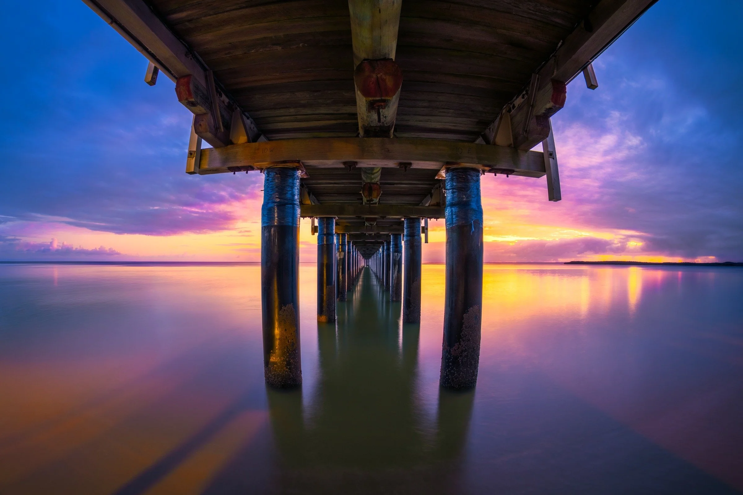 The structure of Urangan Pier stretches across still water beneath a pink and purple evening sky, with reflections drawing the eye towards the horizon.
