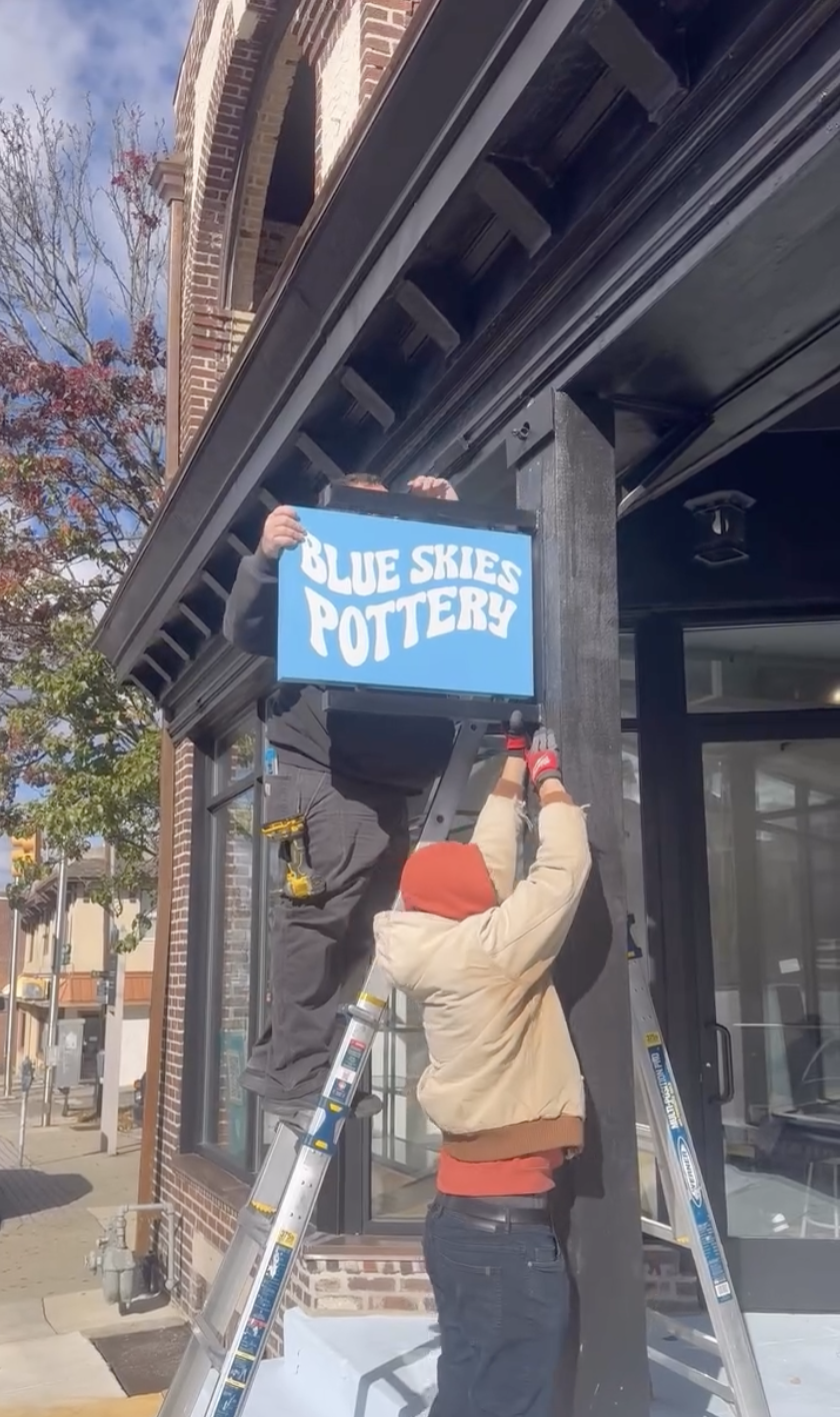 The hanging Blue Skies Pottery sign being installed from a ladder
