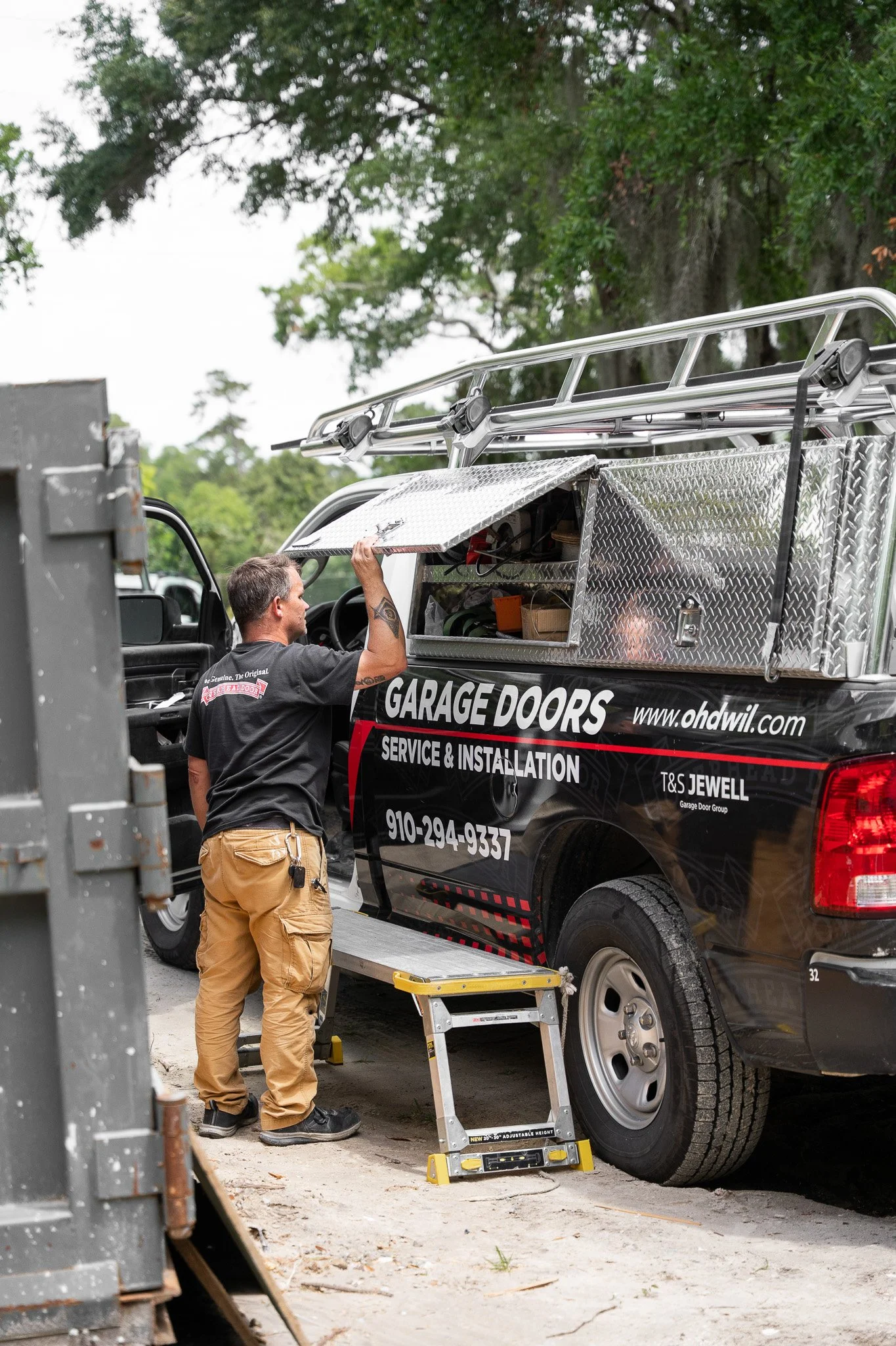 A Overhead Door Company employee working on the side of a service truck labeled 'Garage Doors' parked on a concrete driveway with trees in the background.