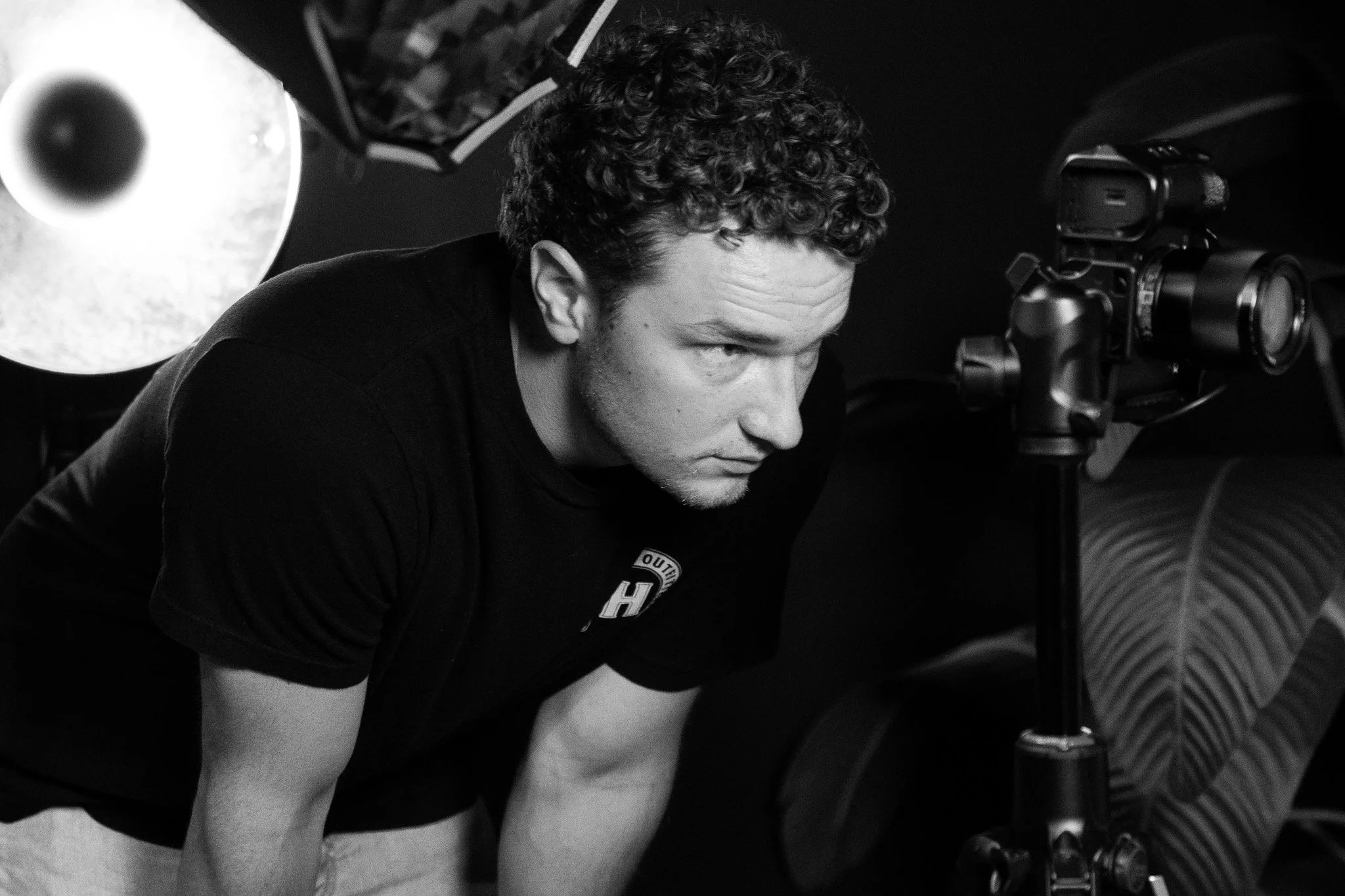 Bradley Gossett is looking intently at a camera mounted on a tripod in a dark setting with studio lighting.
