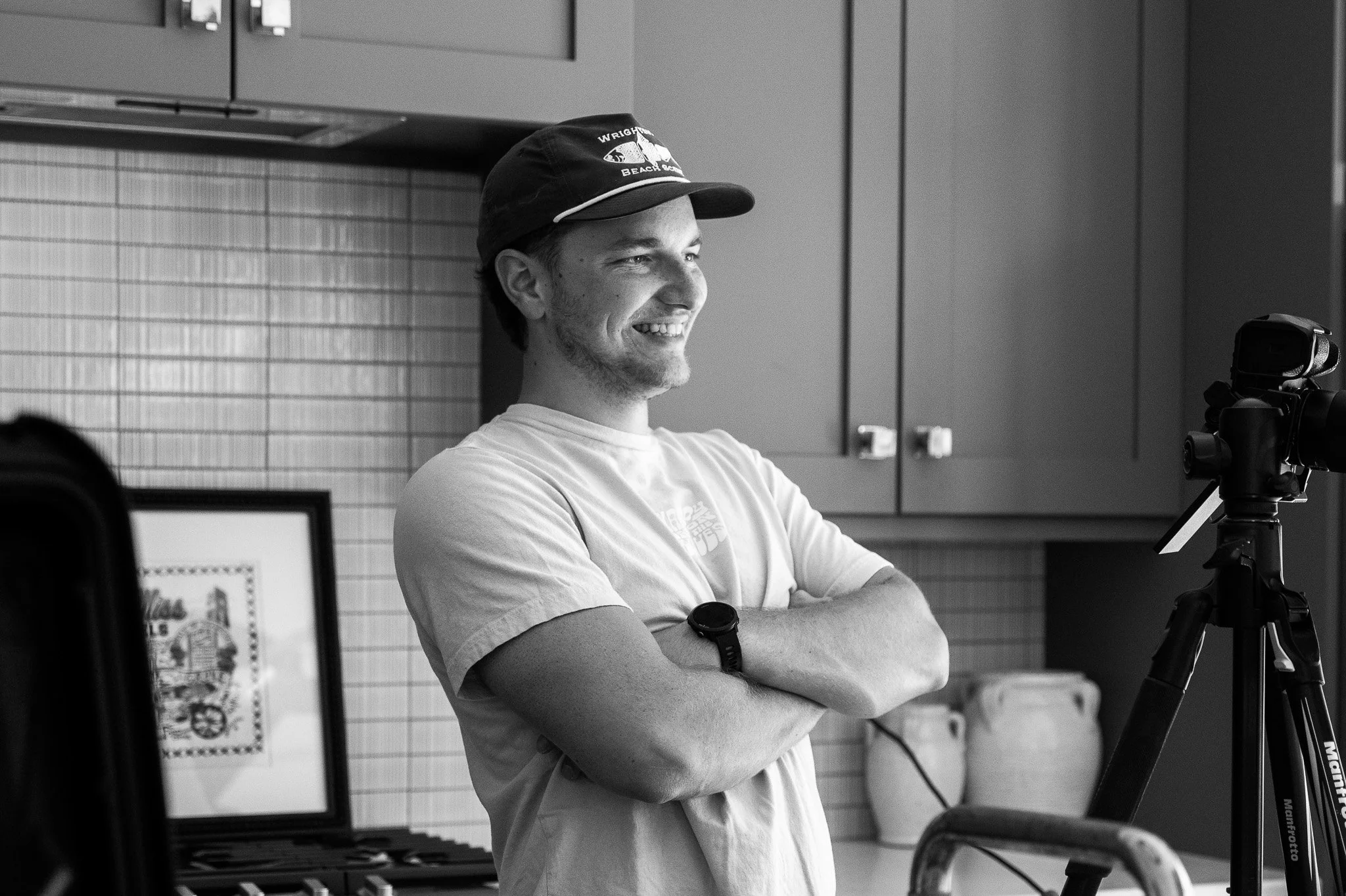 Bradley Gossett, wearing a cap and a t-shirt, is smiling with arms crossed, standing next to a camera on a tripod in a kitchen. The background shows kitchen cabinets, a framed picture, and kitchen jars.
