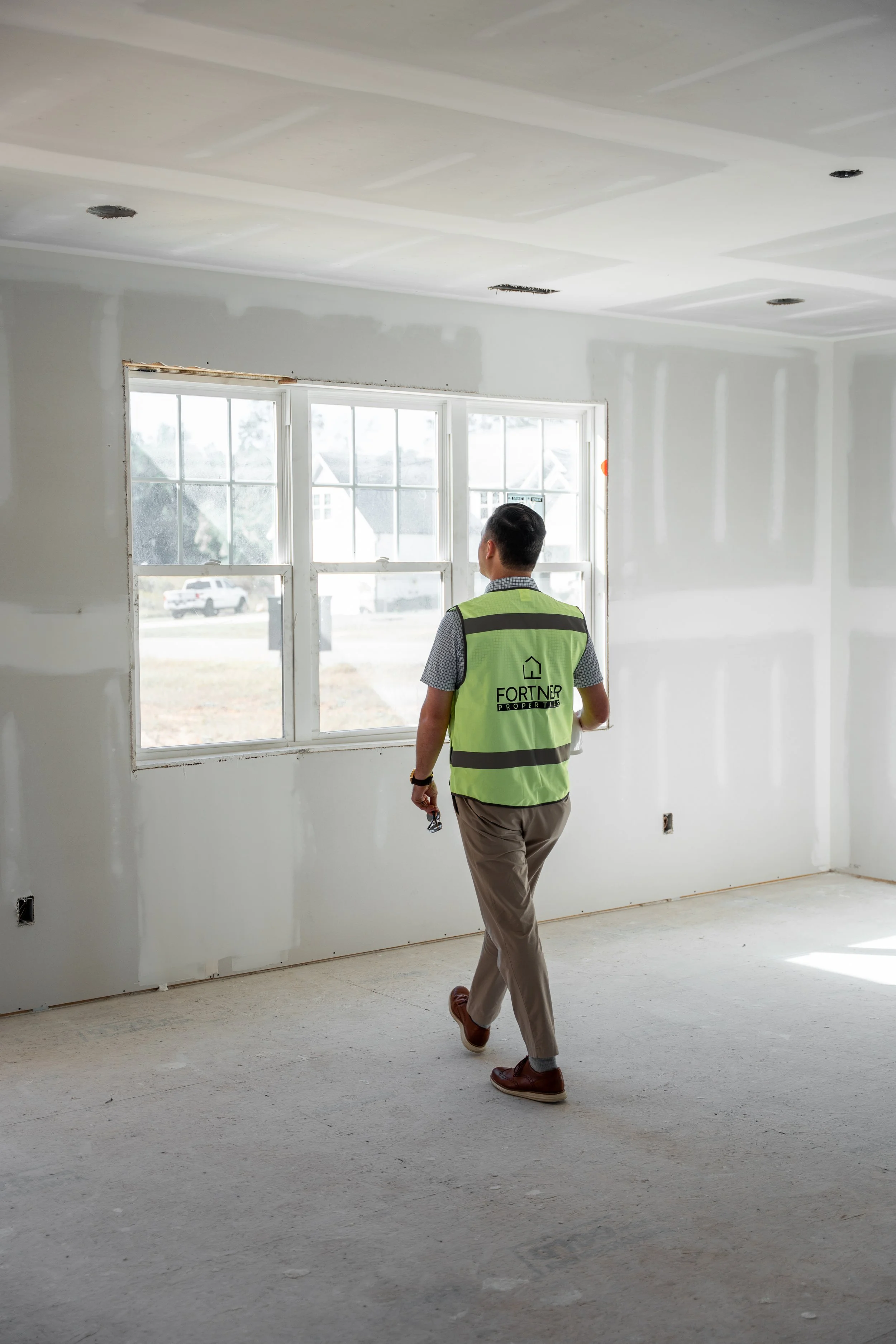 The owner of Fortner Homes with yellow safety vest inspecting an unfinished room with drywall, windows, and electrical outlets.