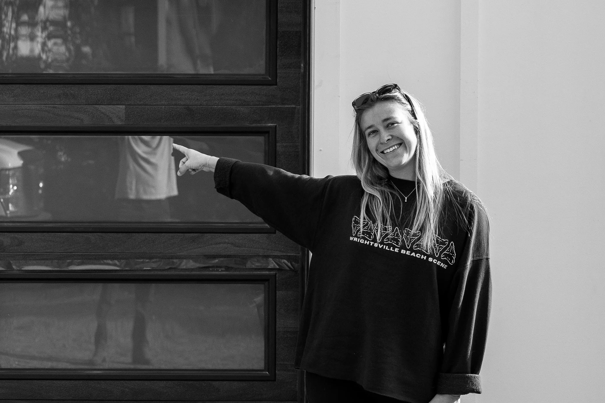 Faith Harris, wearing a black sweatshirt that says 'Wrightsville Beach Scene,' is smiling and pointing at a display case or panel on the wall next to her.